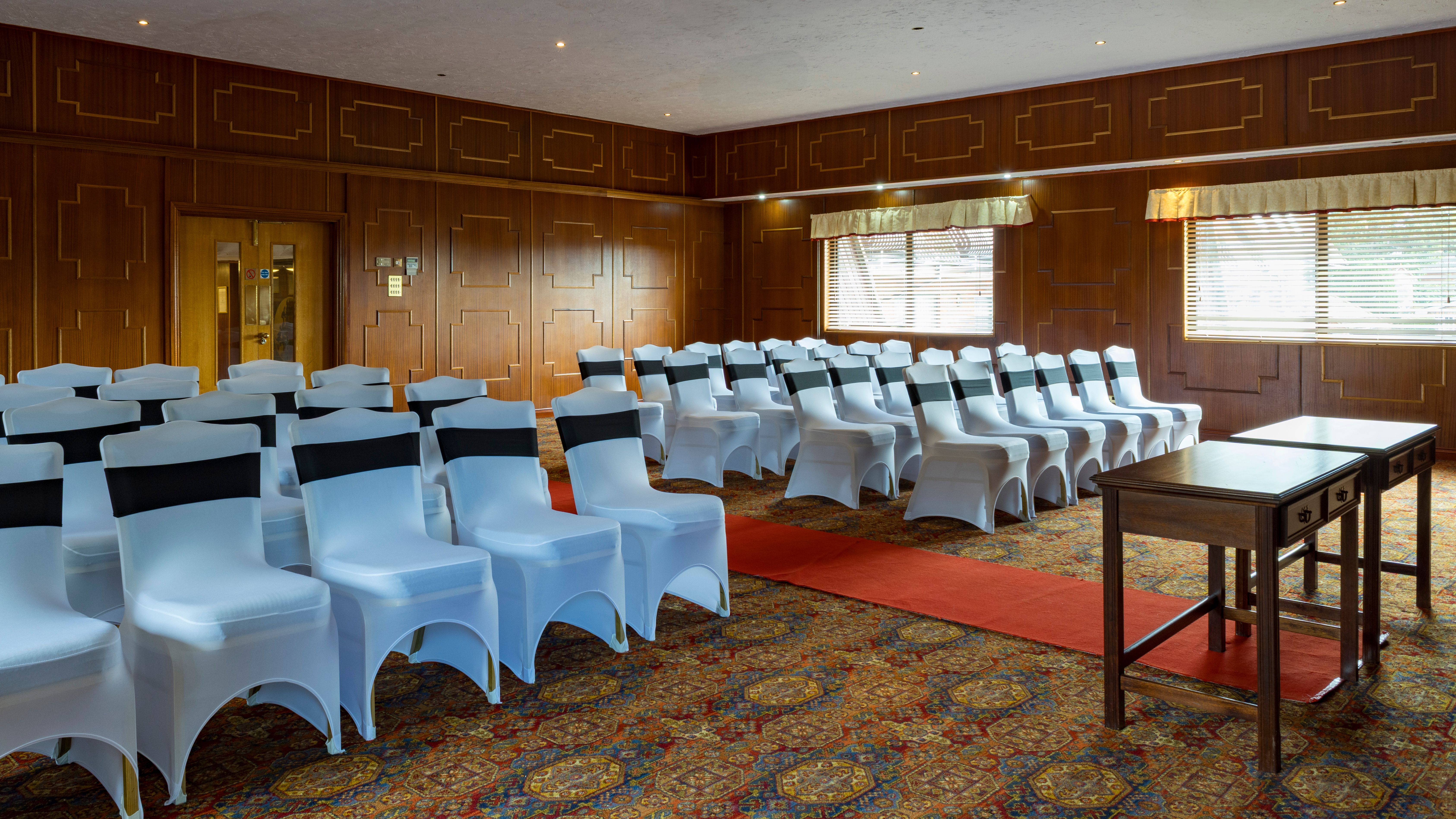 Rows of white chairs with black sashes arranged in a decorated room with wooden panel walls and a red carpet, facing two wooden tables.