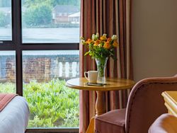 Cozy room corner with a round wooden table, vase of flowers, and cup near a window overlooking greenery and water.