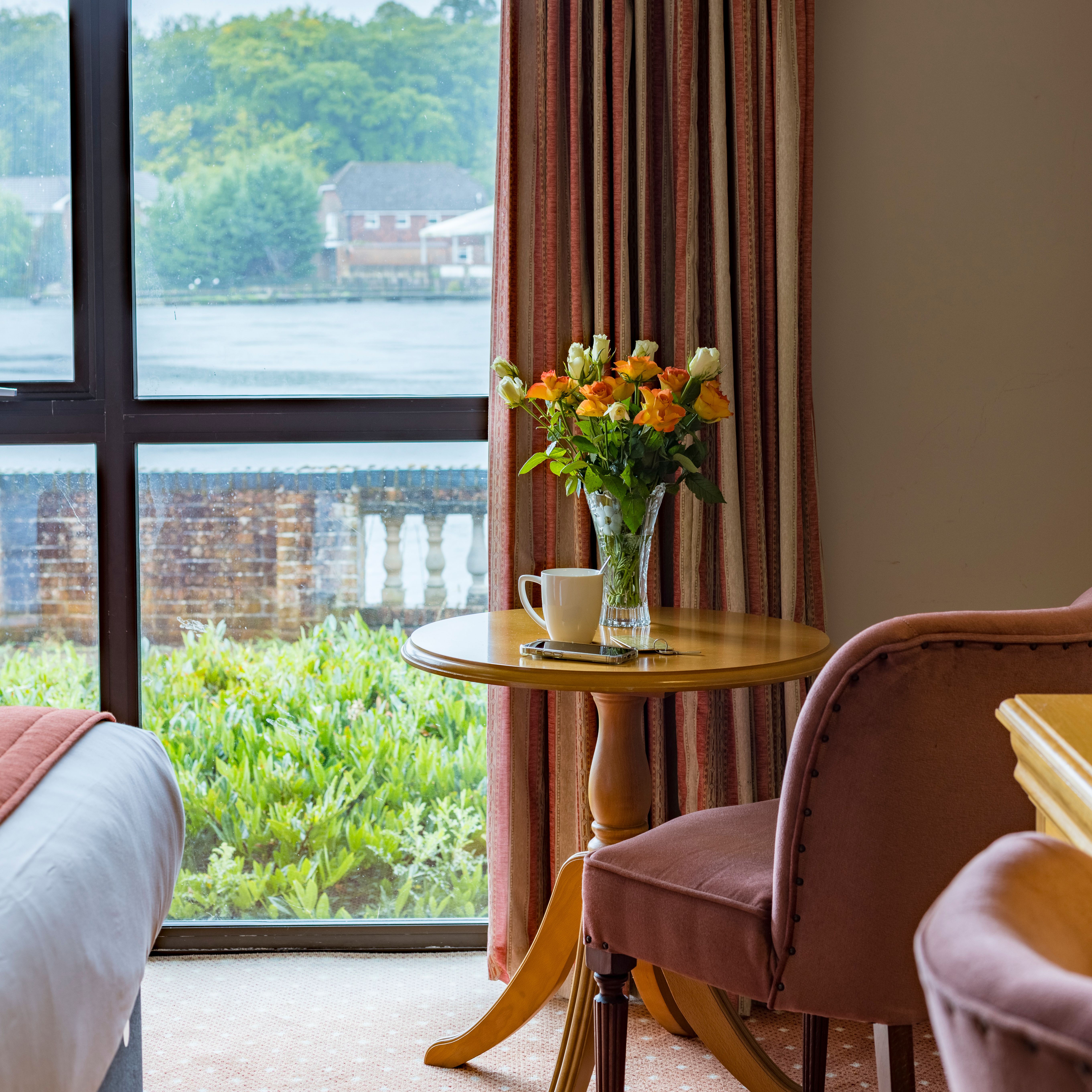 Cozy room corner with a round wooden table, vase of flowers, and cup near a window overlooking greenery and water.