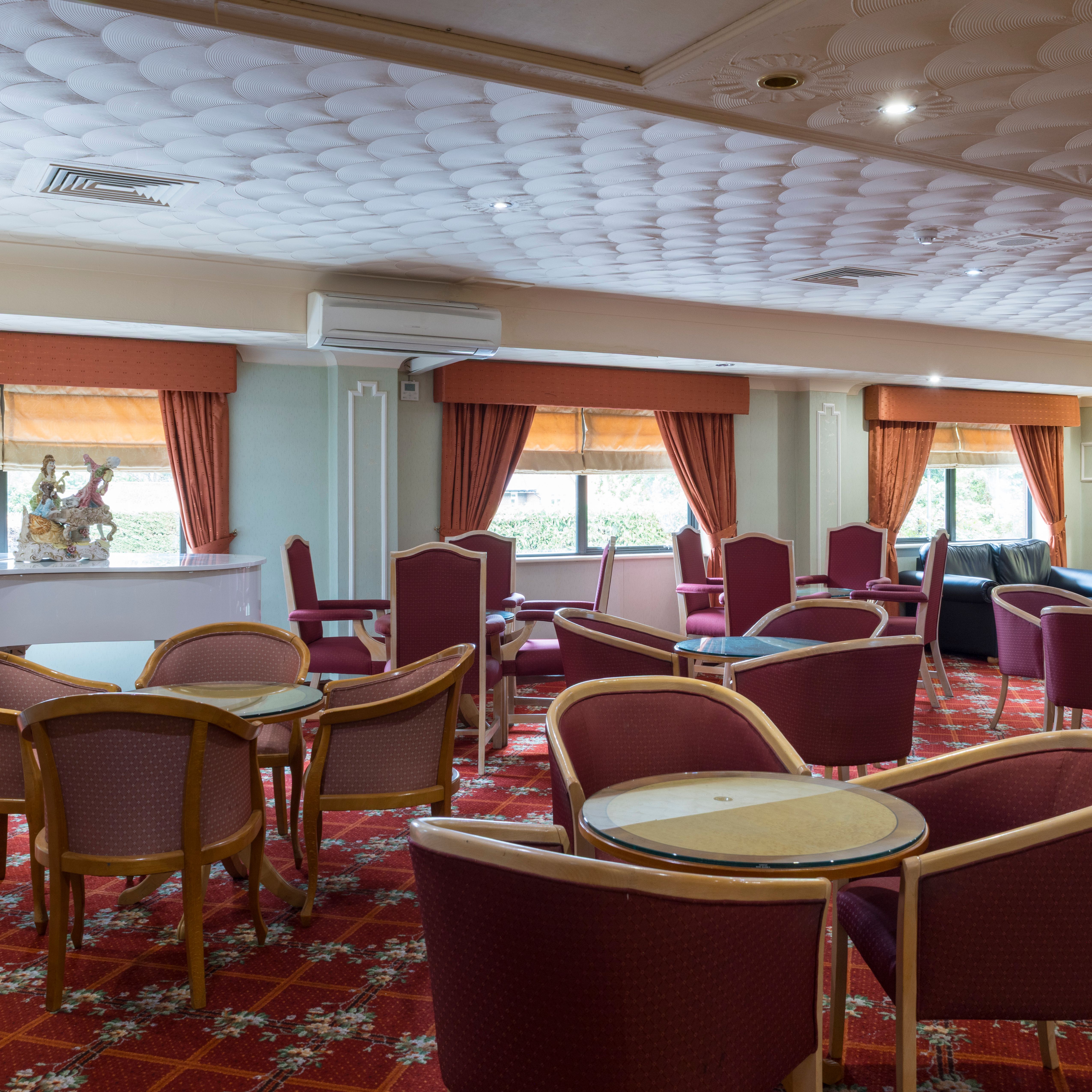 Spacious lounge with red patterned carpet, round tables, red chairs, white piano, and large windows with orange curtains.