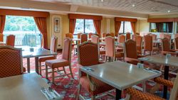 Empty hotel restaurant with orange chairs, set tables, and large windows with curtains overlooking a lake.