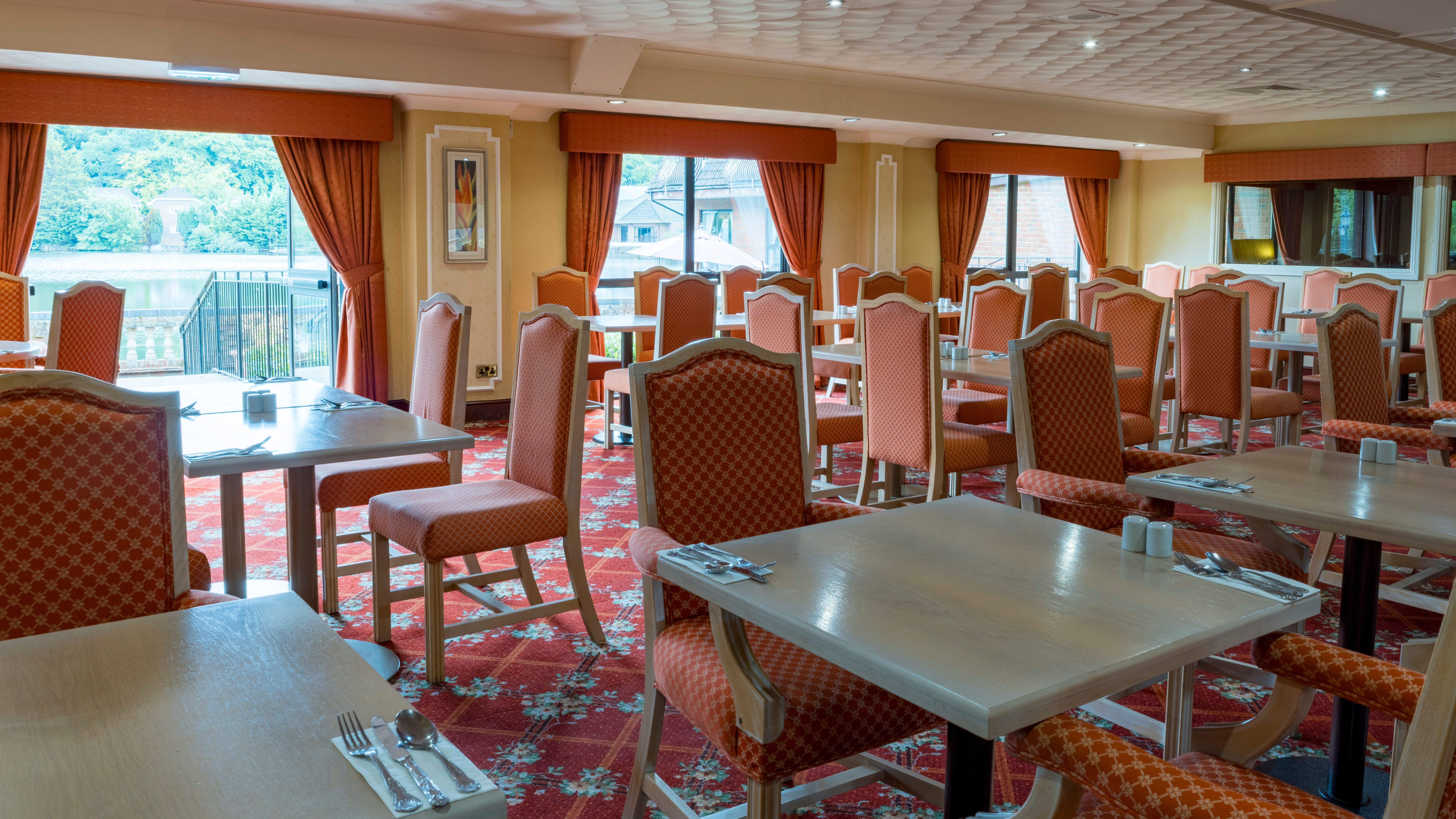 Empty hotel restaurant with orange chairs, set tables, and large windows with curtains overlooking a lake.