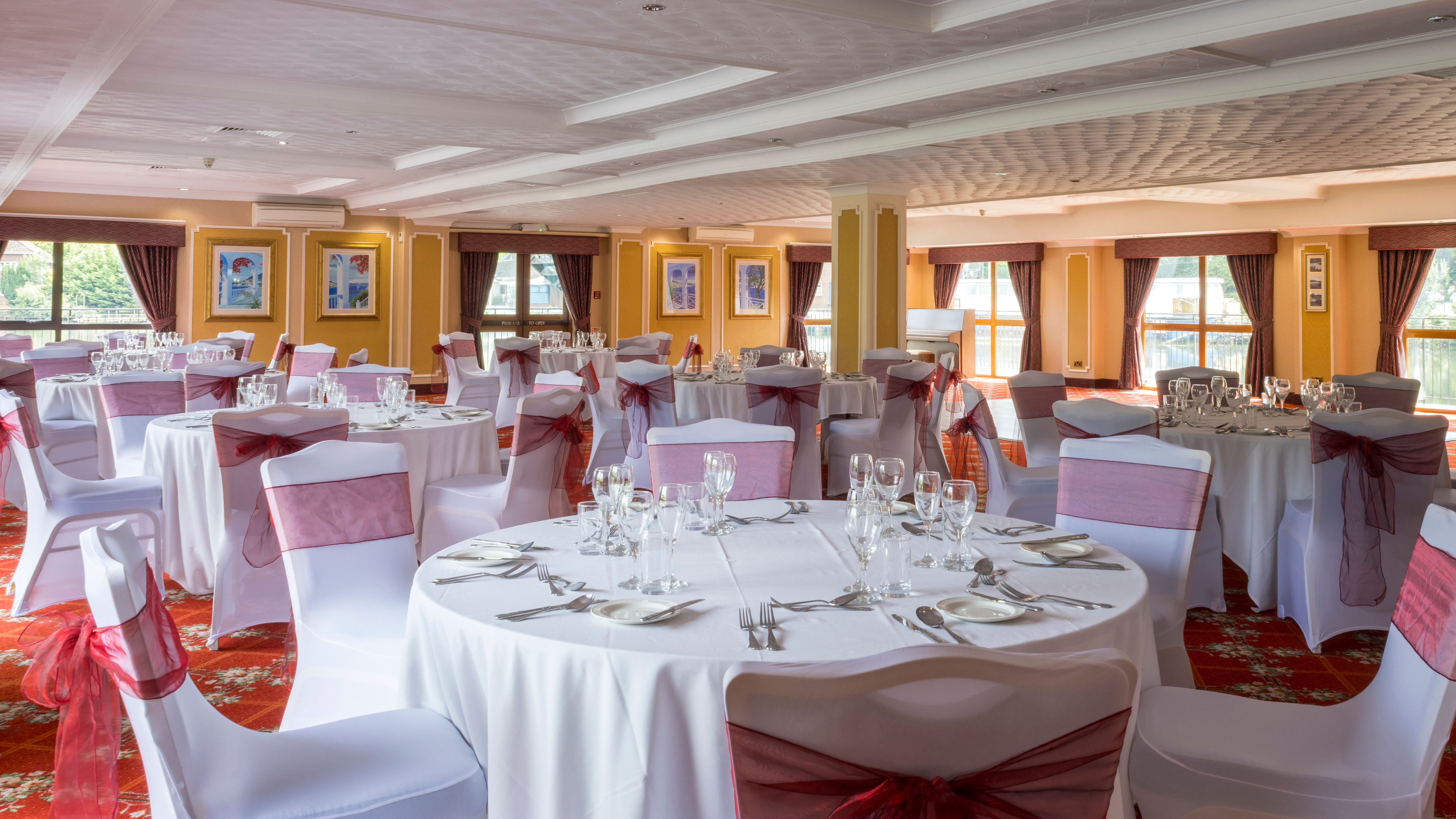 Elegant banquet hall with round tables set for an event, featuring white tablecloths and chairs with burgundy bows.