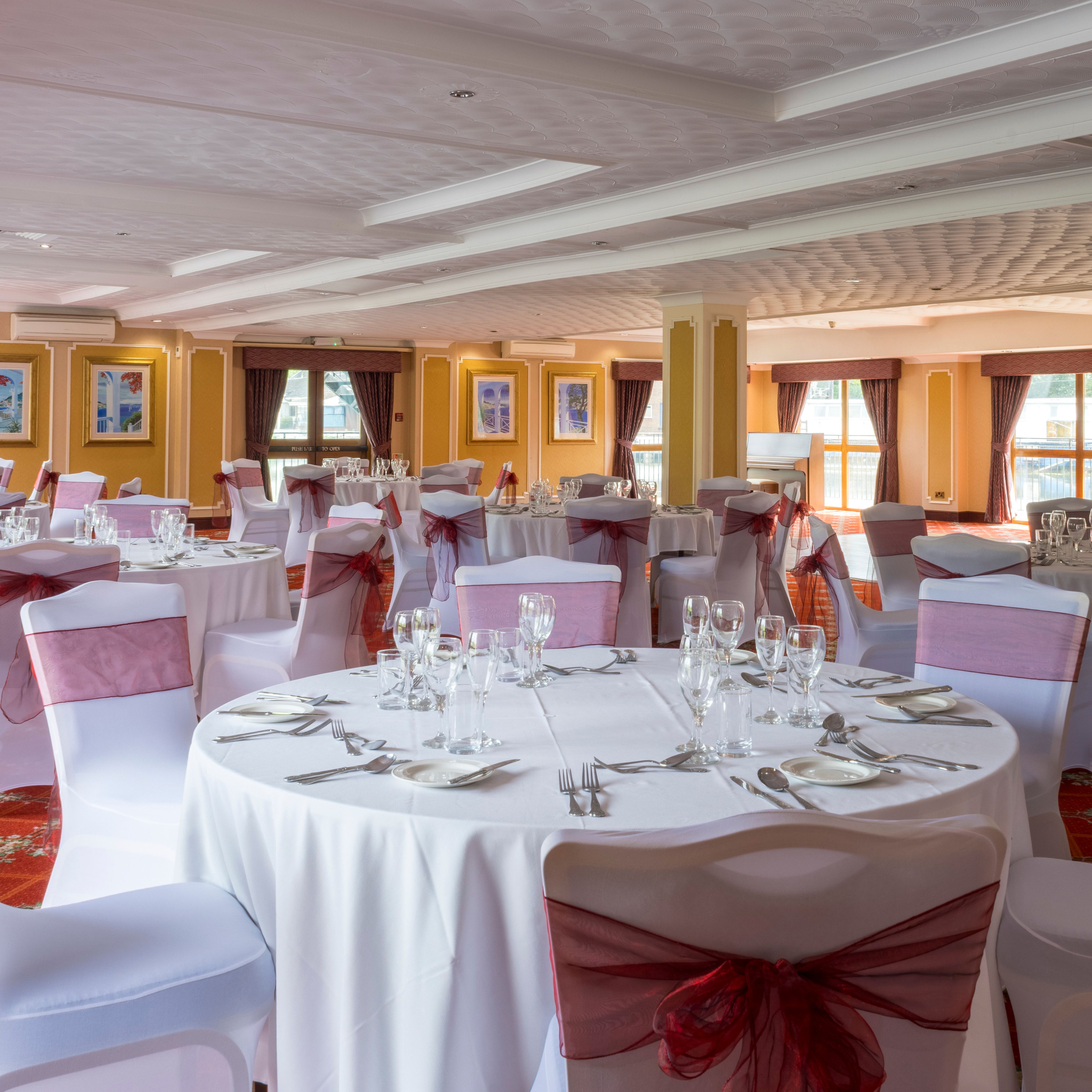 Elegant banquet hall with round tables set for an event, featuring white tablecloths and chairs with burgundy bows.