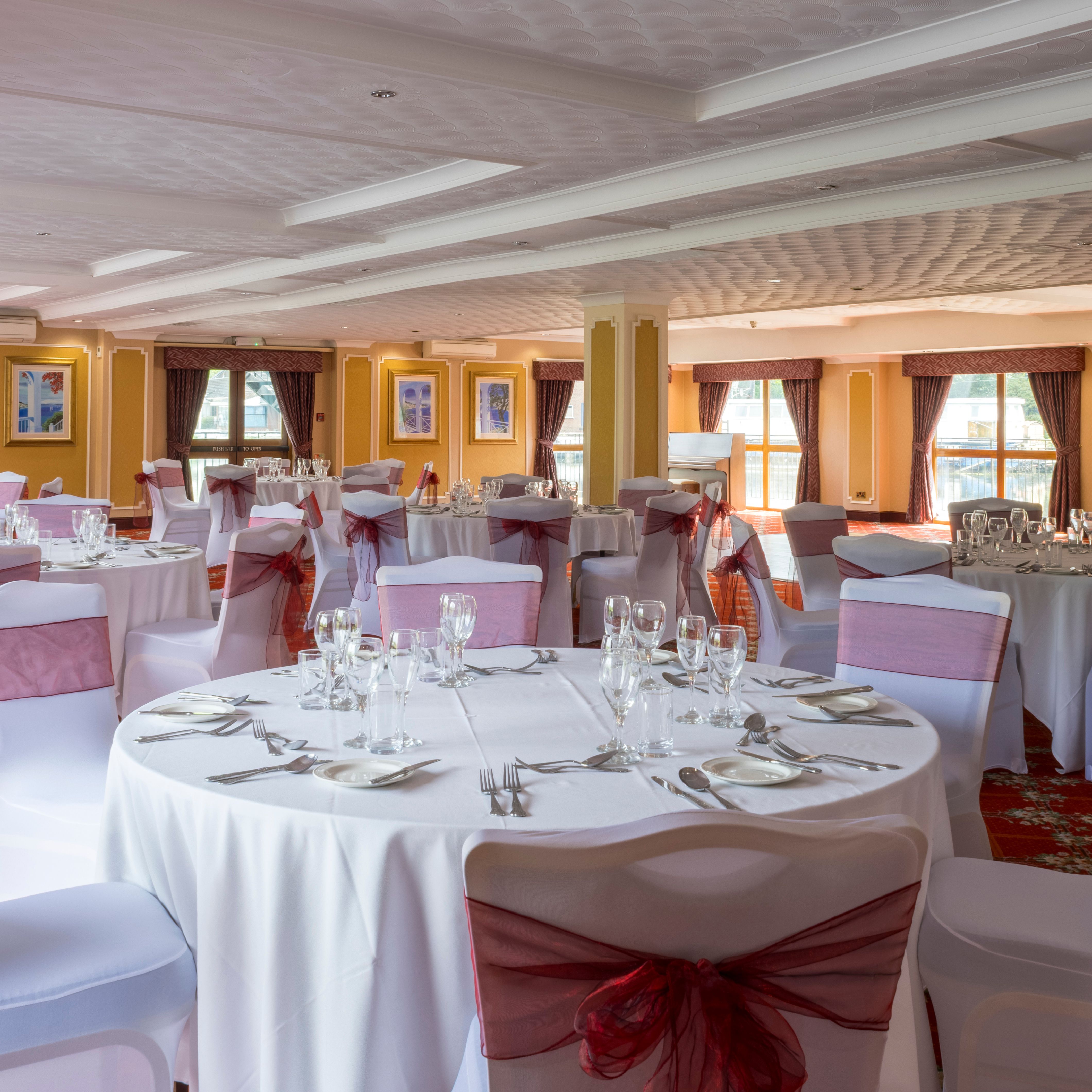 Elegant banquet hall with round tables set for an event, featuring white tablecloths and chairs with burgundy bows.