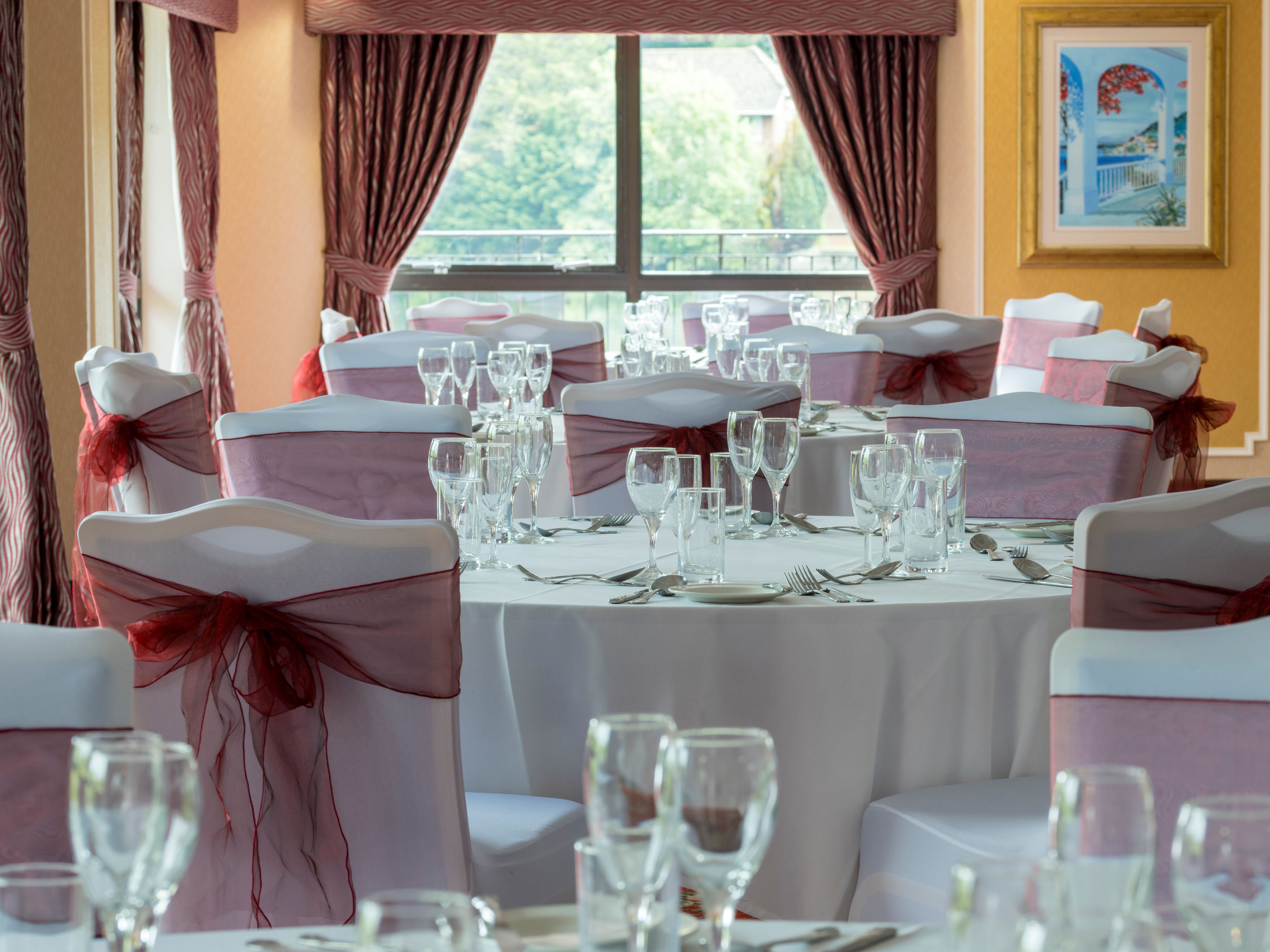 Elegant banquet hall with round tables set for an event, featuring white tablecloths, wine glasses, and chairs decorated with burgundy sashes.