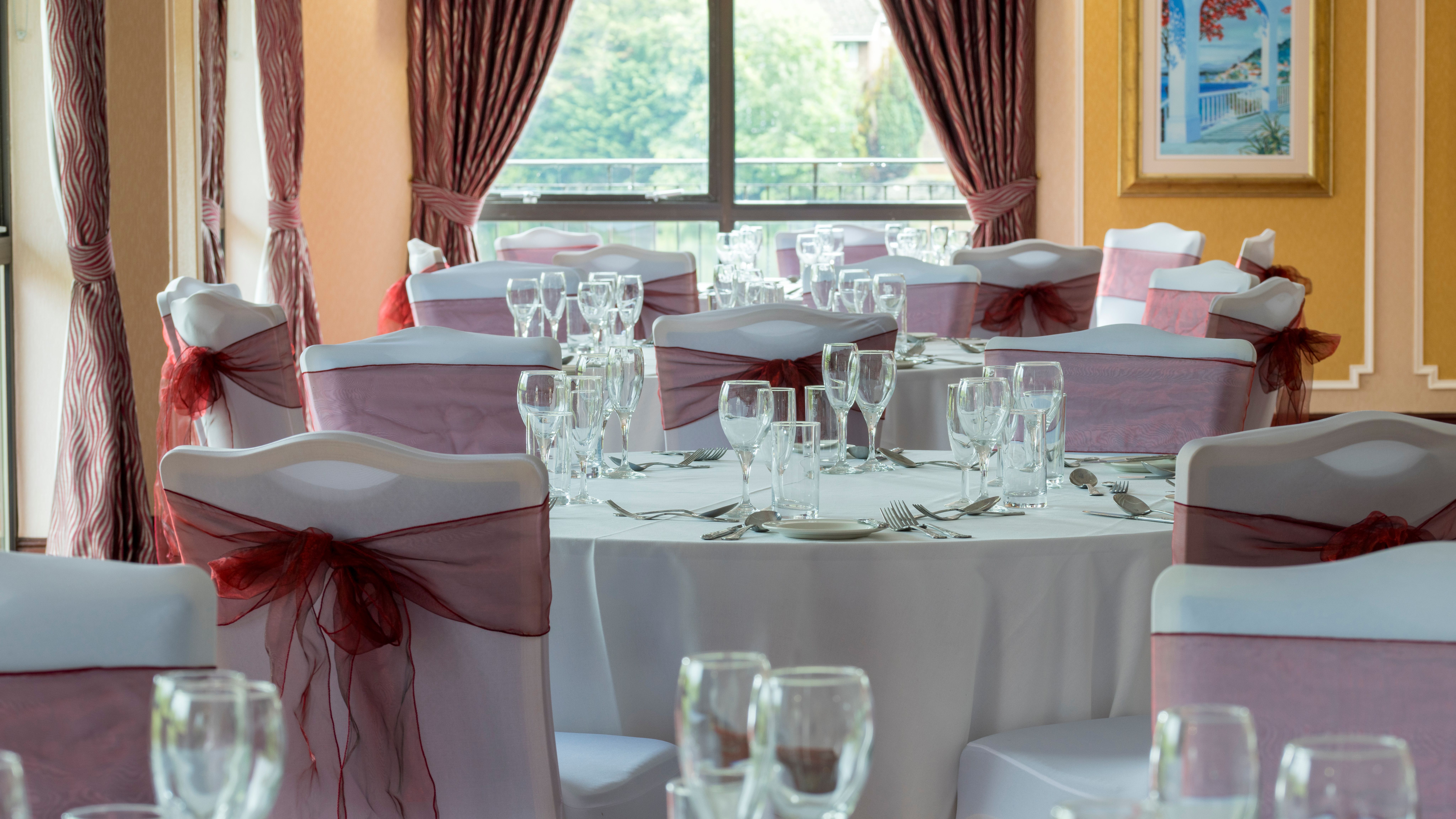 Elegant banquet hall with round tables set for an event, featuring white tablecloths, wine glasses, and chairs decorated with burgundy sashes.
