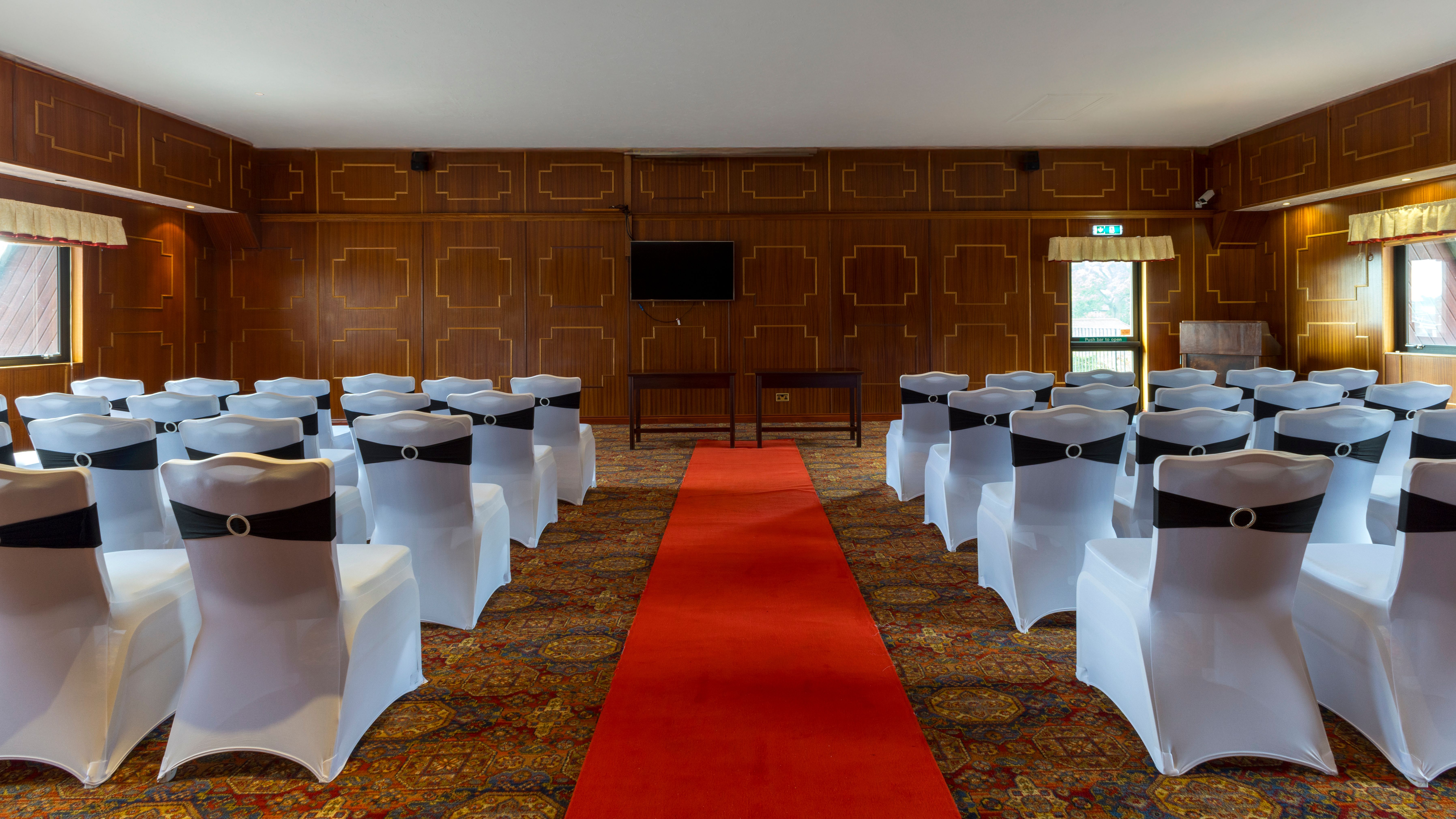 Room set up for an event with rows of white chairs, a red carpet, and wood-paneled walls.