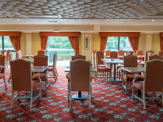 Spacious dining room with red patterned carpet, orange chairs, and large windows with orange curtains overlooking a scenic outdoor view.
