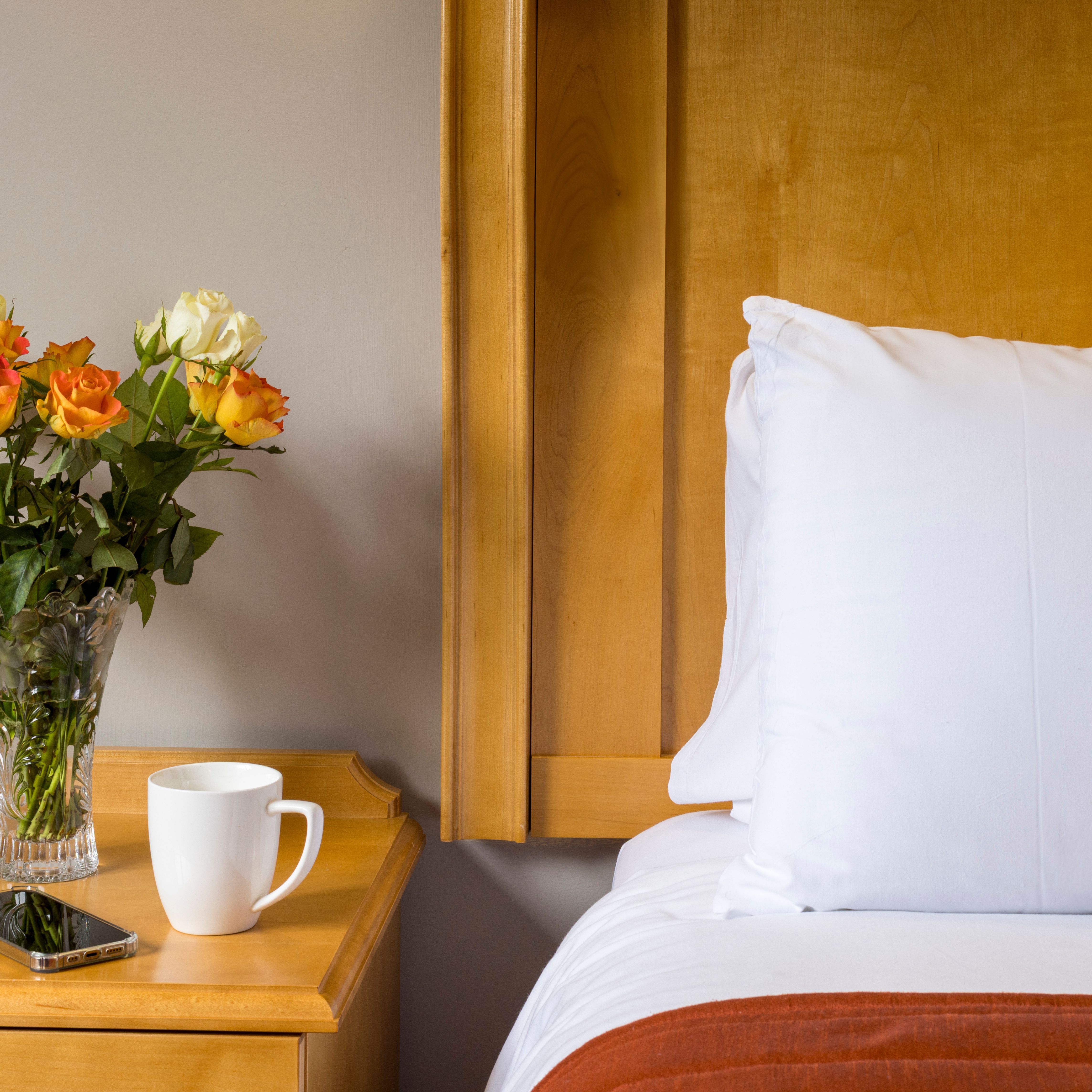 A bedside table with a vase of orange and white roses, a white coffee mug, and a smartphone next to a bed with white linens.