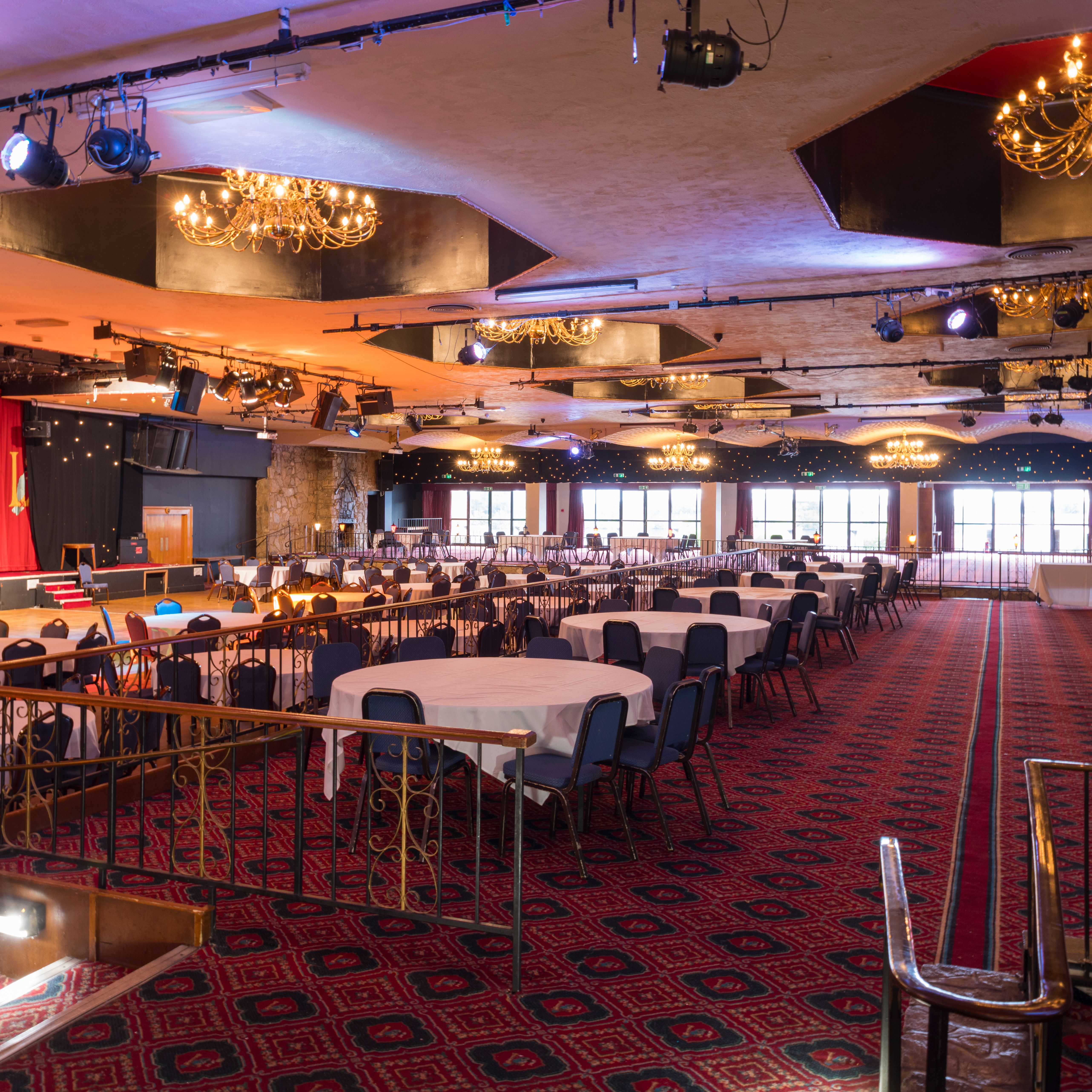 Large banquet hall with round tables and chairs, chandeliers, stage, and red patterned carpet