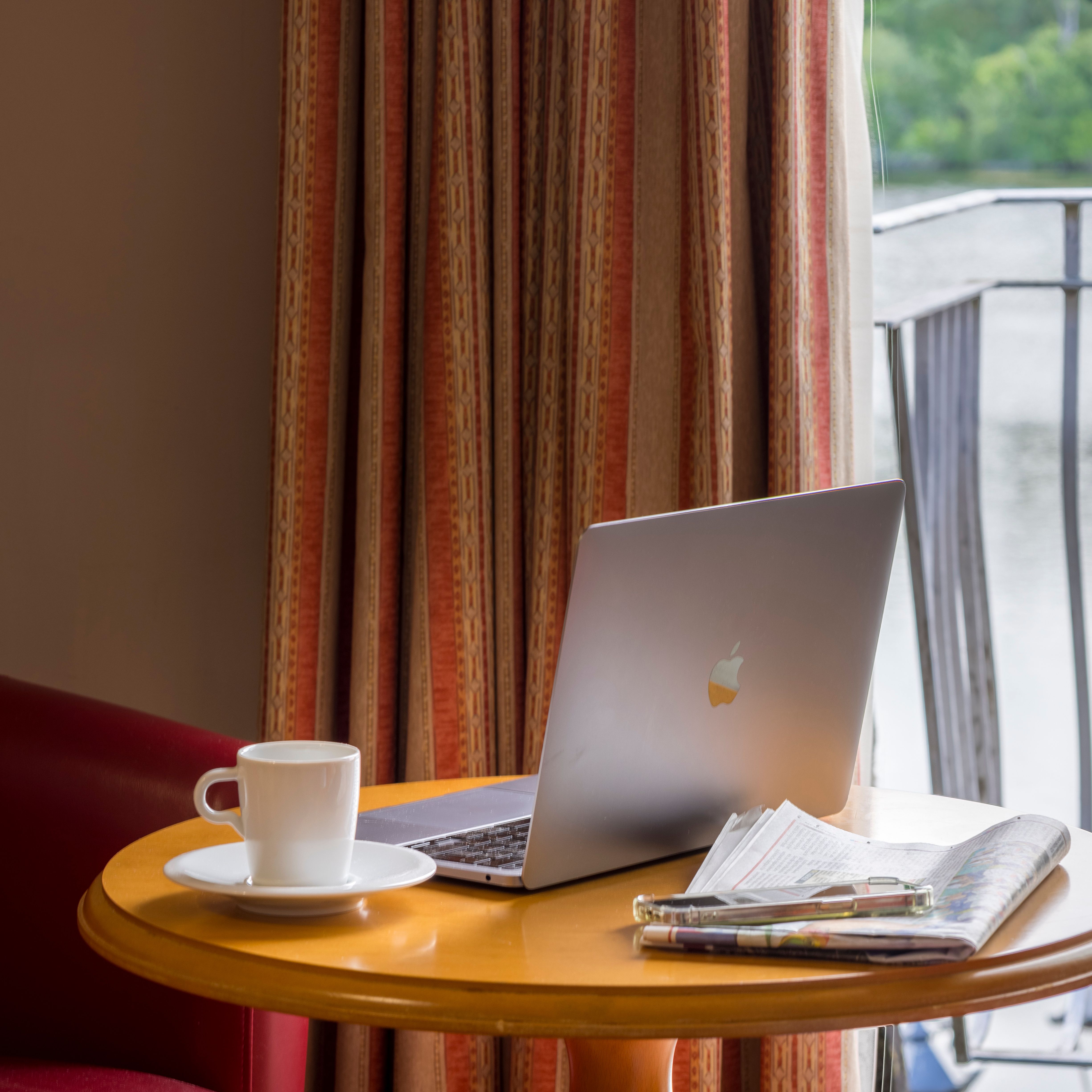 Laptop, cup of coffee, and newspaper on a round table by a window with a view of a lake.