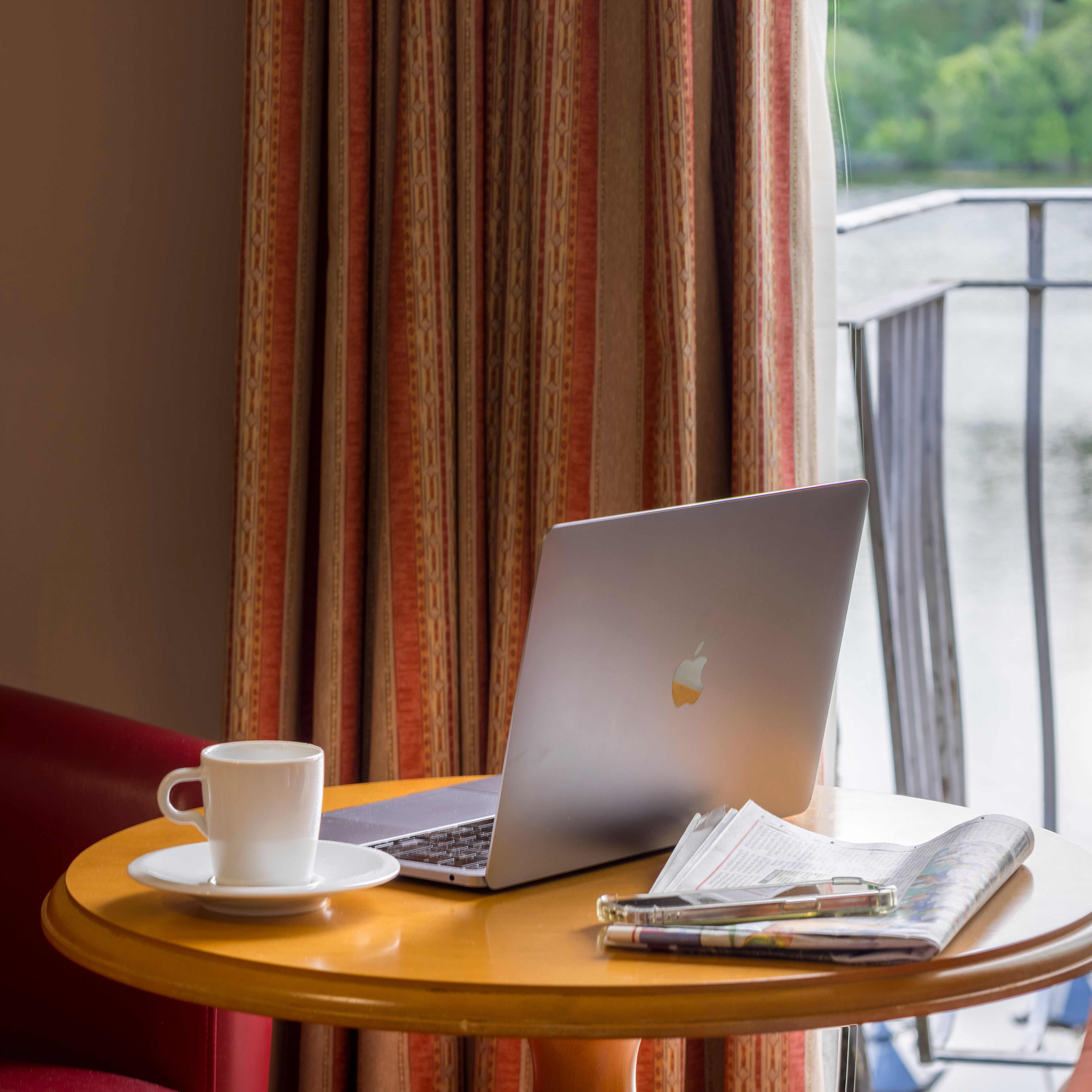 Laptop, cup of coffee, and newspaper on a round table by a window with a view of a lake.