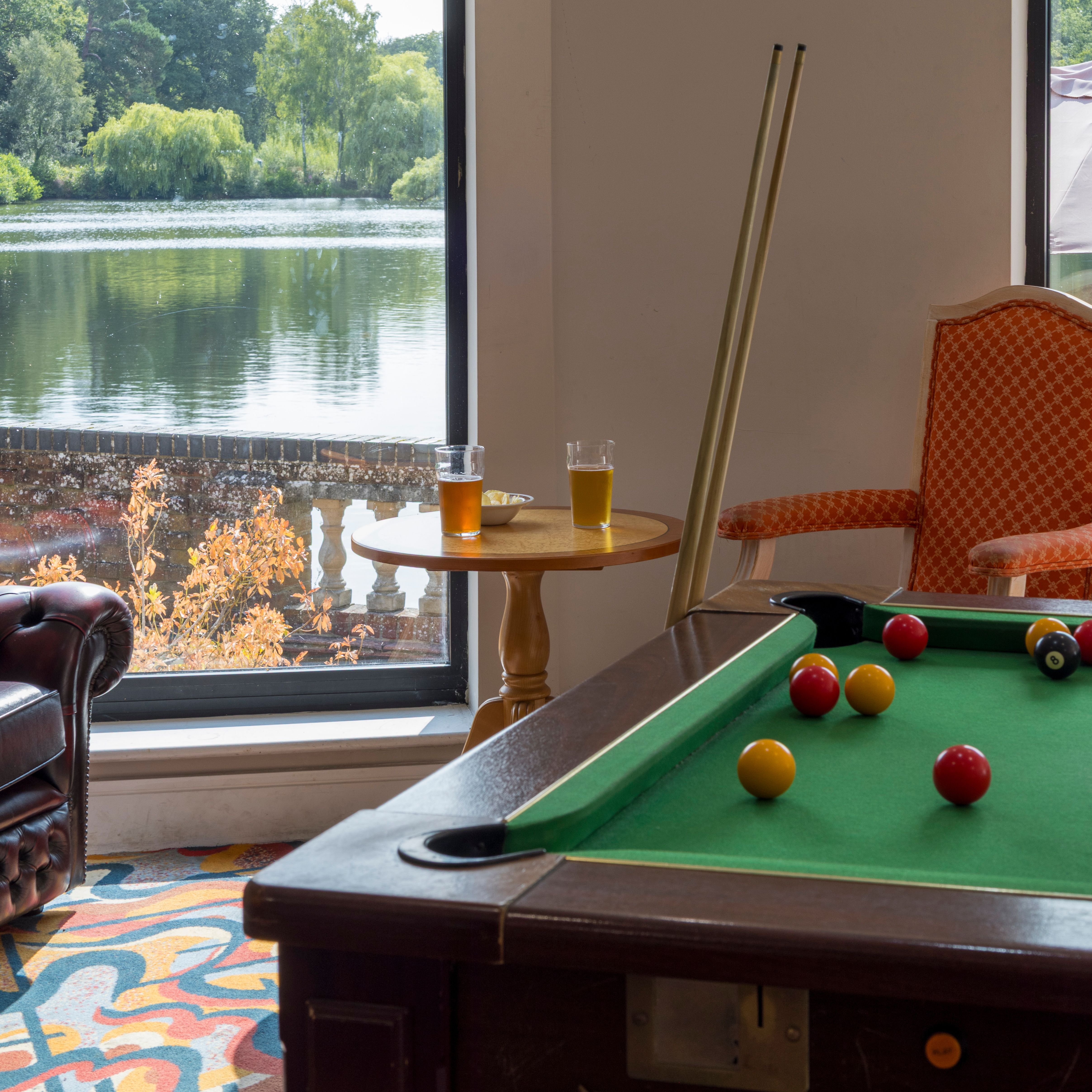 A room with a pool table and large windows overlooking a lake with trees in the background.