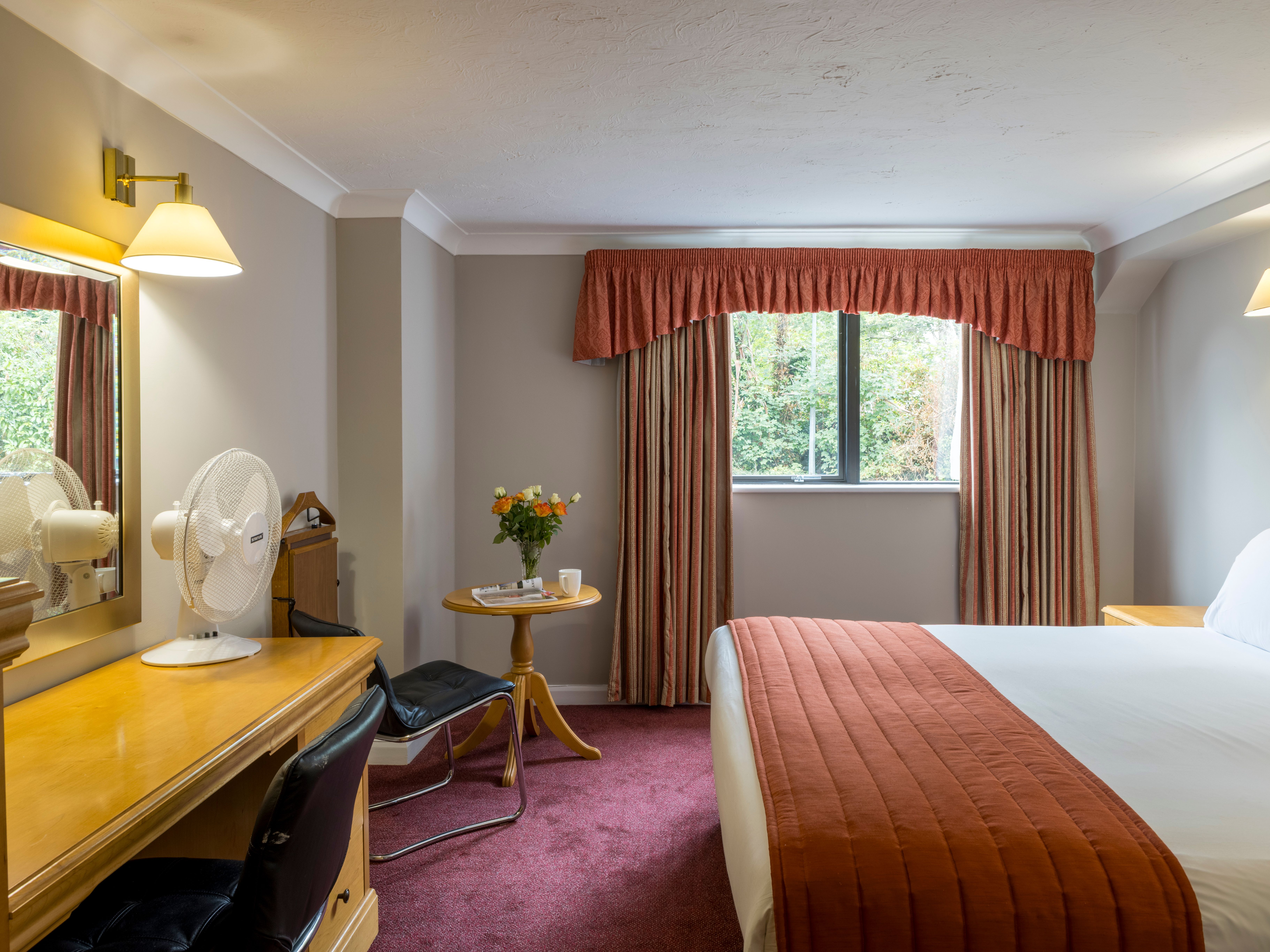 A hotel room with a double bed, desk, chair, window with curtains, and a small table with flowers.