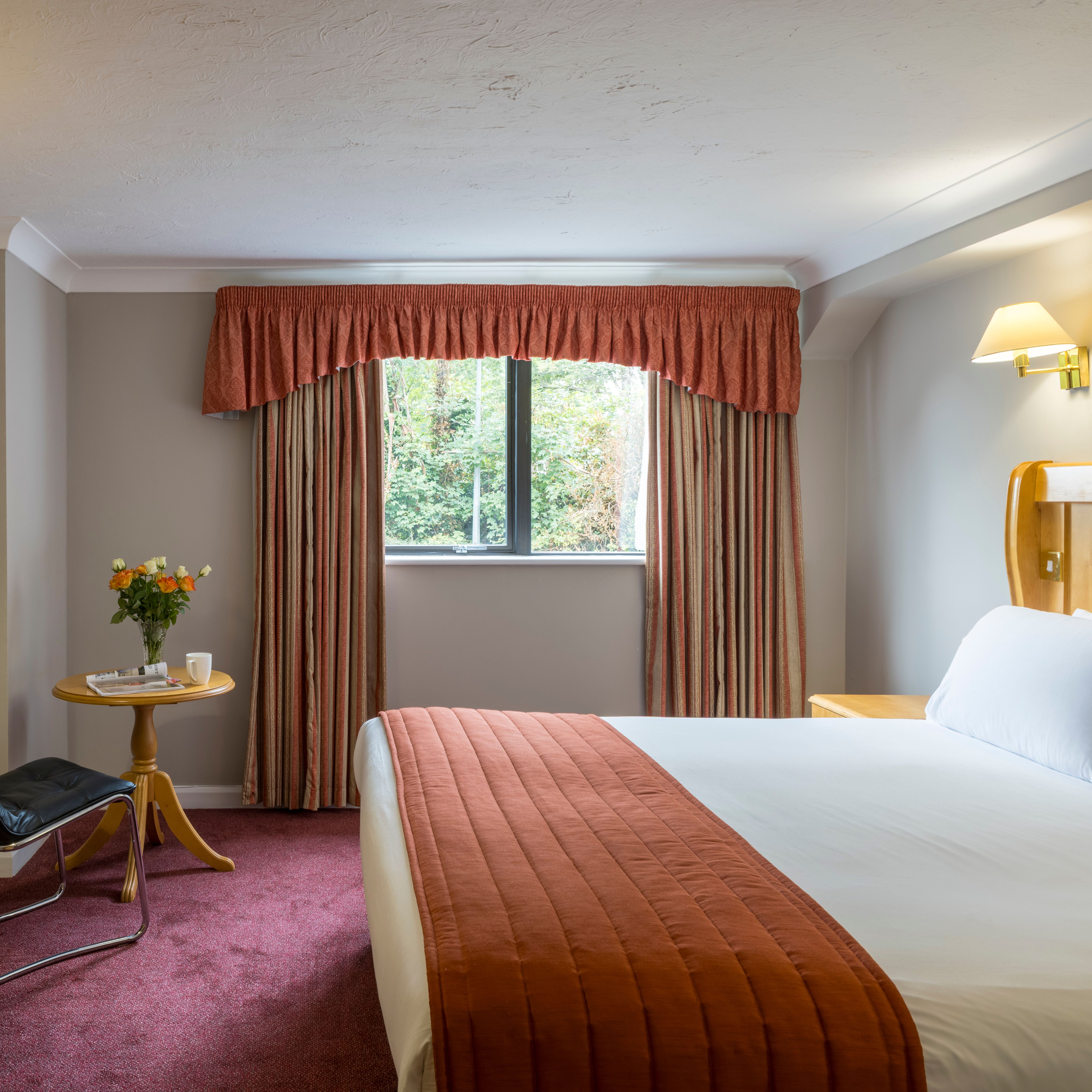 A hotel room with a double bed, desk, chair, window with curtains, and a small table with flowers.
