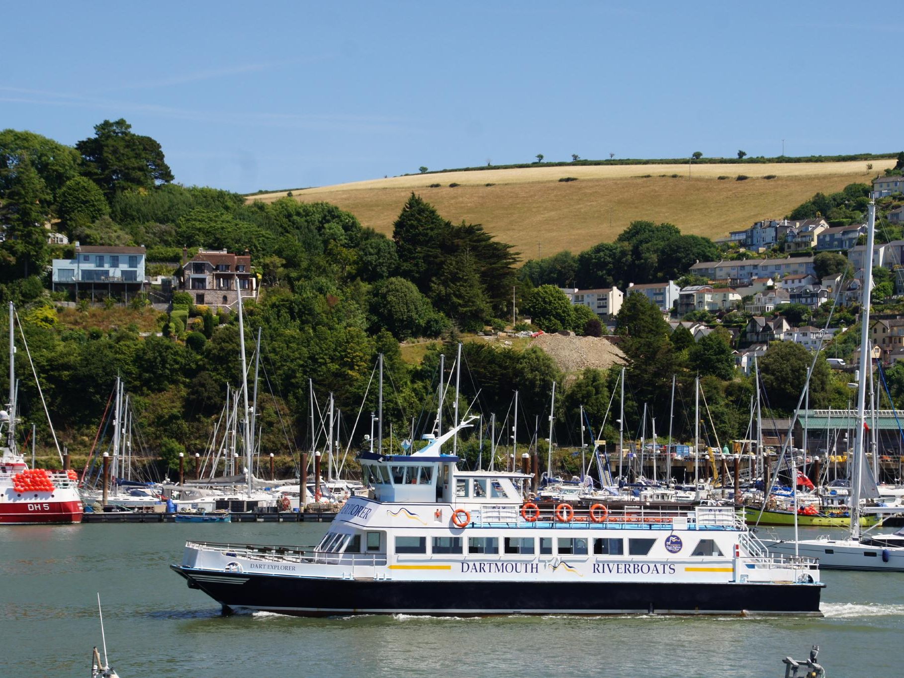 Passenger ferry passes a marina with sailing boats, with hillside houses and fields above the river in Dartmouth, Devon.