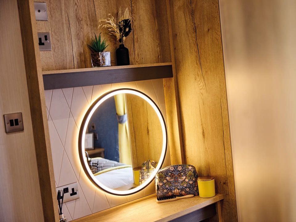 Round illuminated mirror above a wooden dressing table with toiletry bag and candle