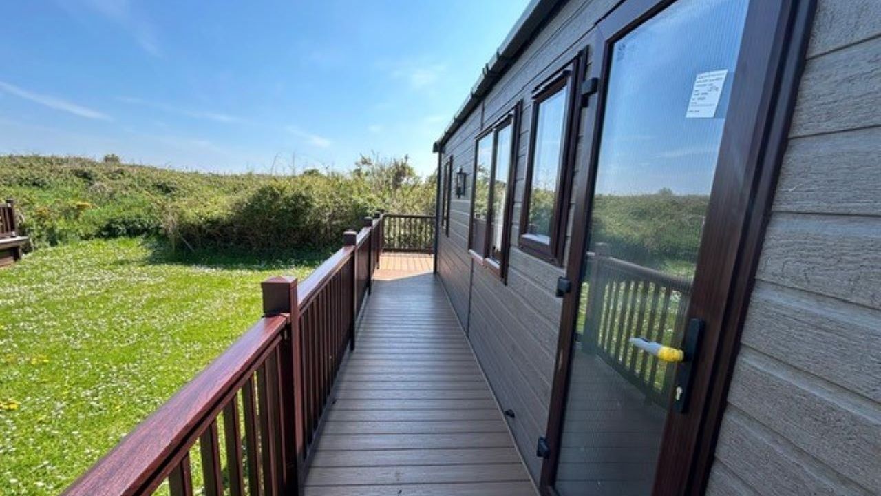 Wooden decking walkway alongside a grey static caravan with railing and glass door, overlooking a grassy field