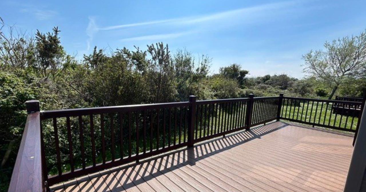 Timber deck with dark railing overlooking green shrubs and trees under a blue sky