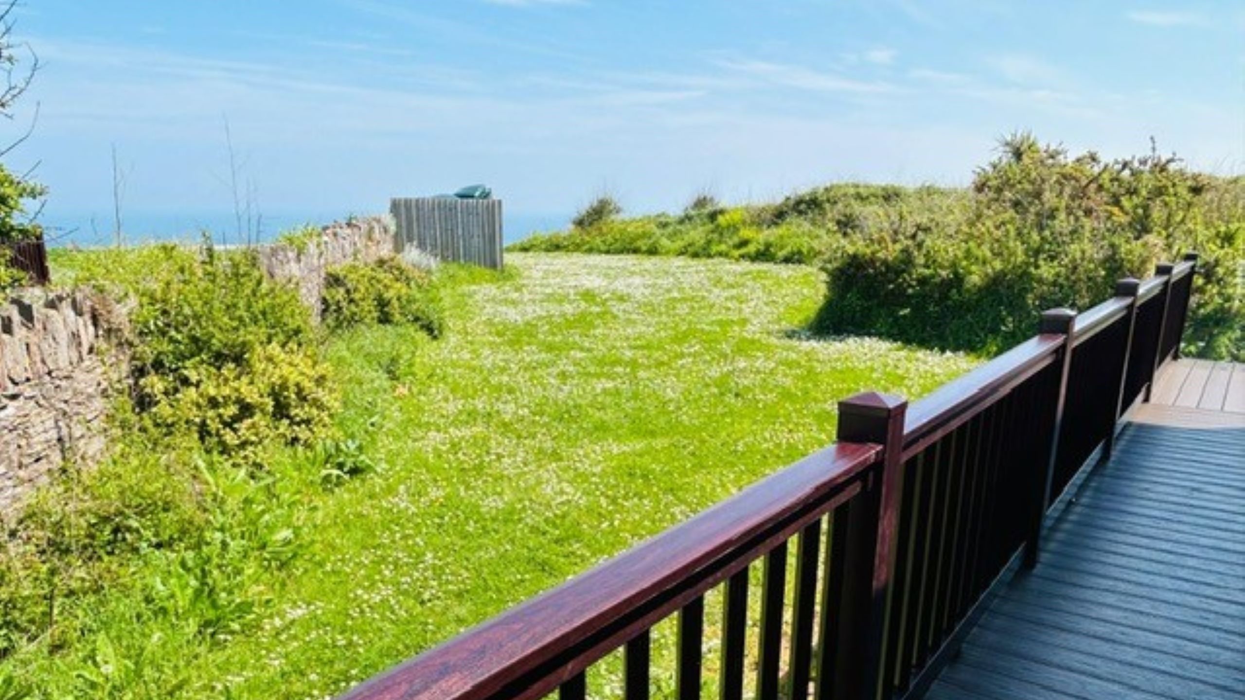 Wooden deck with dark railing overlooking a sunny grassy garden with coastal views