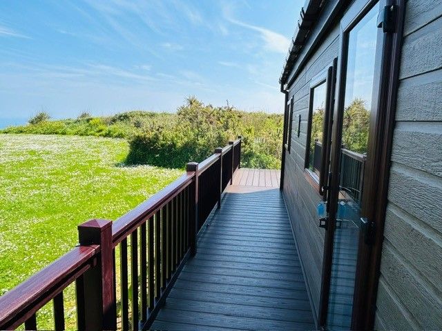 Wooden deck walkway beside a lodge with railings, overlooking a grassy field under a blue sky