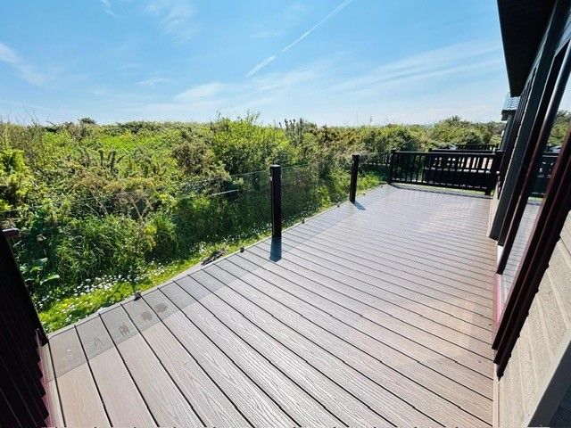 Sunlit wooden decking with glass balustrade overlooking green shrubs under a blue sky