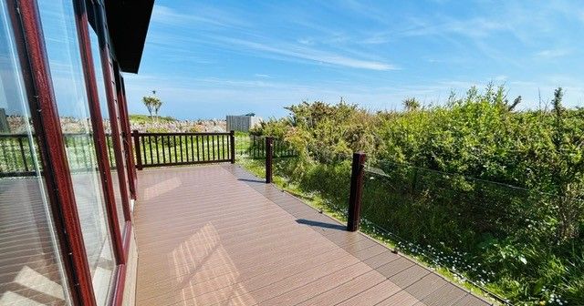 Wooden decked terrace beside a glass-fronted building, overlooking coastal greenery and blue sky