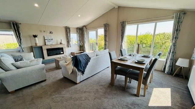 Bright open-plan lounge and dining area with grey sofas, dining table and large windows overlooking greenery