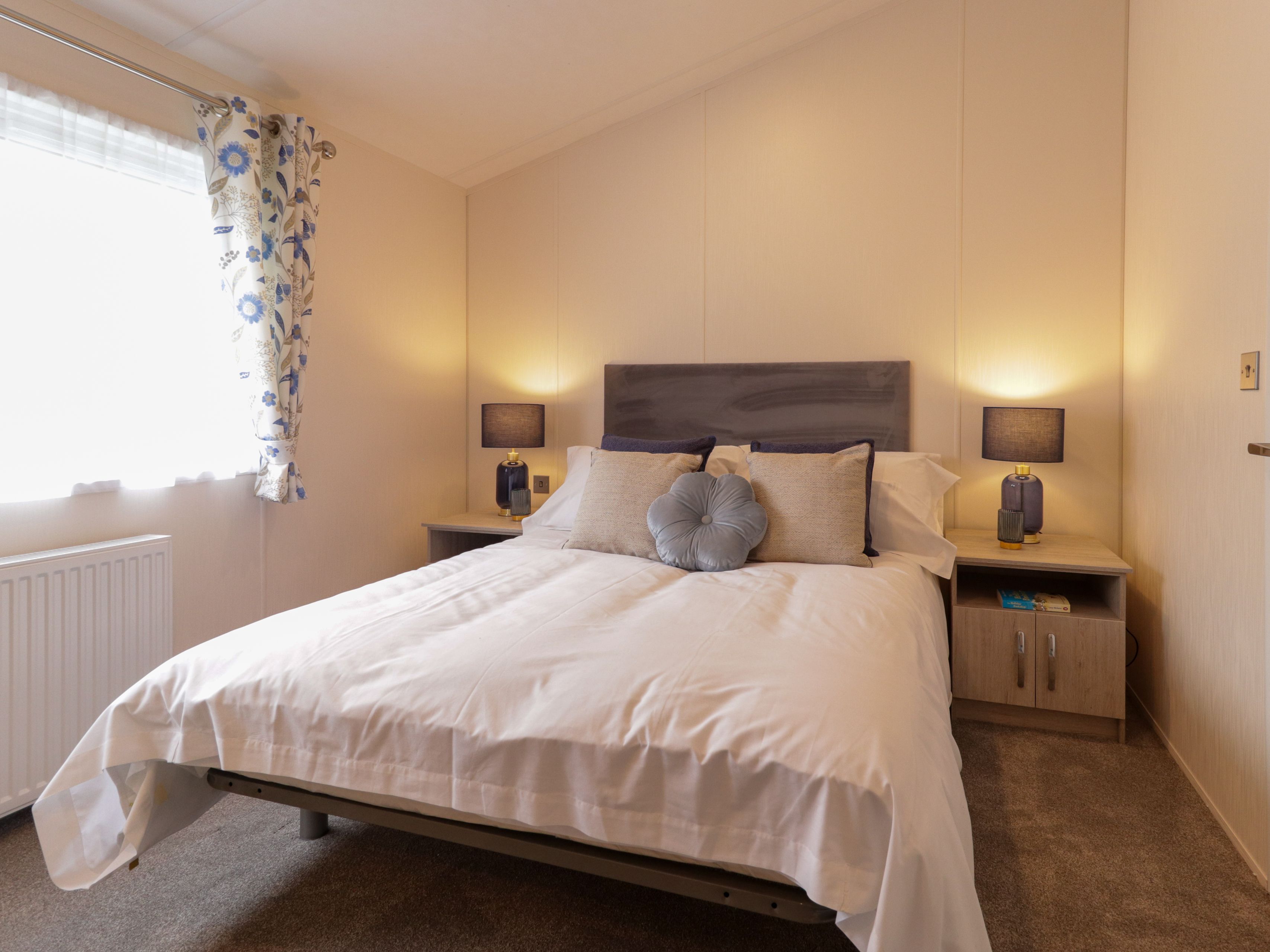 Cosy bedroom with double bed, grey headboard, bedside tables and lamps, and a window with floral curtains