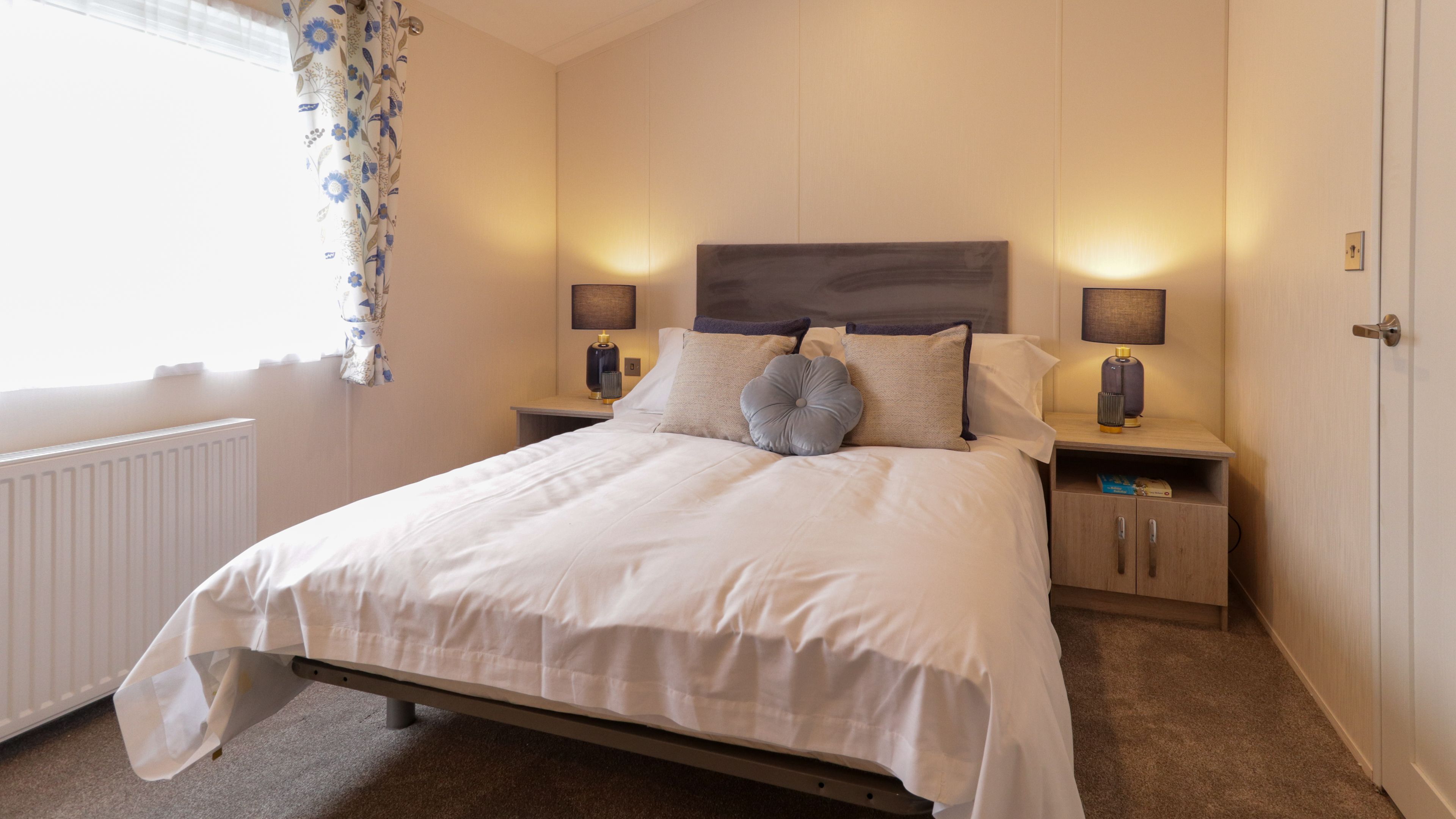 Cosy bedroom with double bed, grey headboard, bedside tables and lamps, and a window with floral curtains
