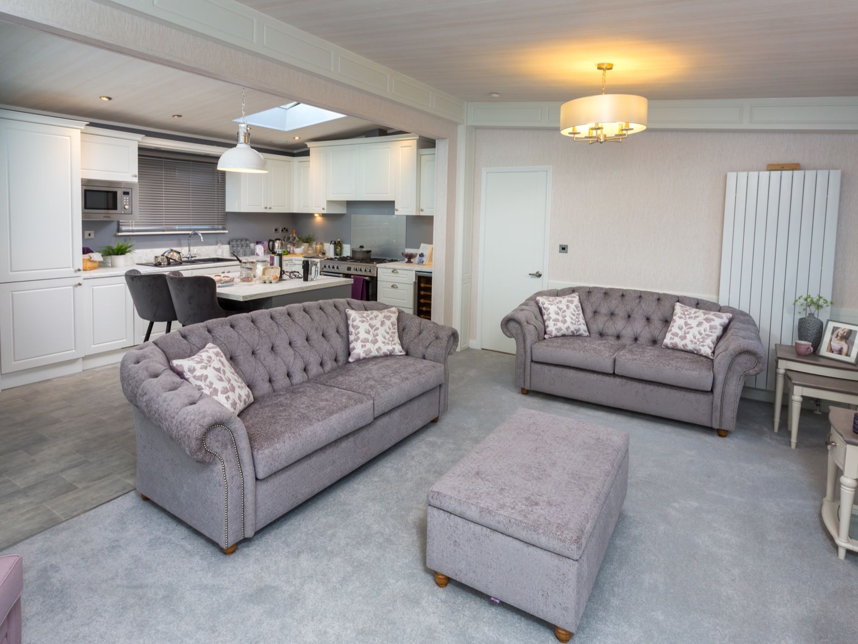 Open-plan living room and kitchen with grey tufted sofas, ottoman, and white cabinetry