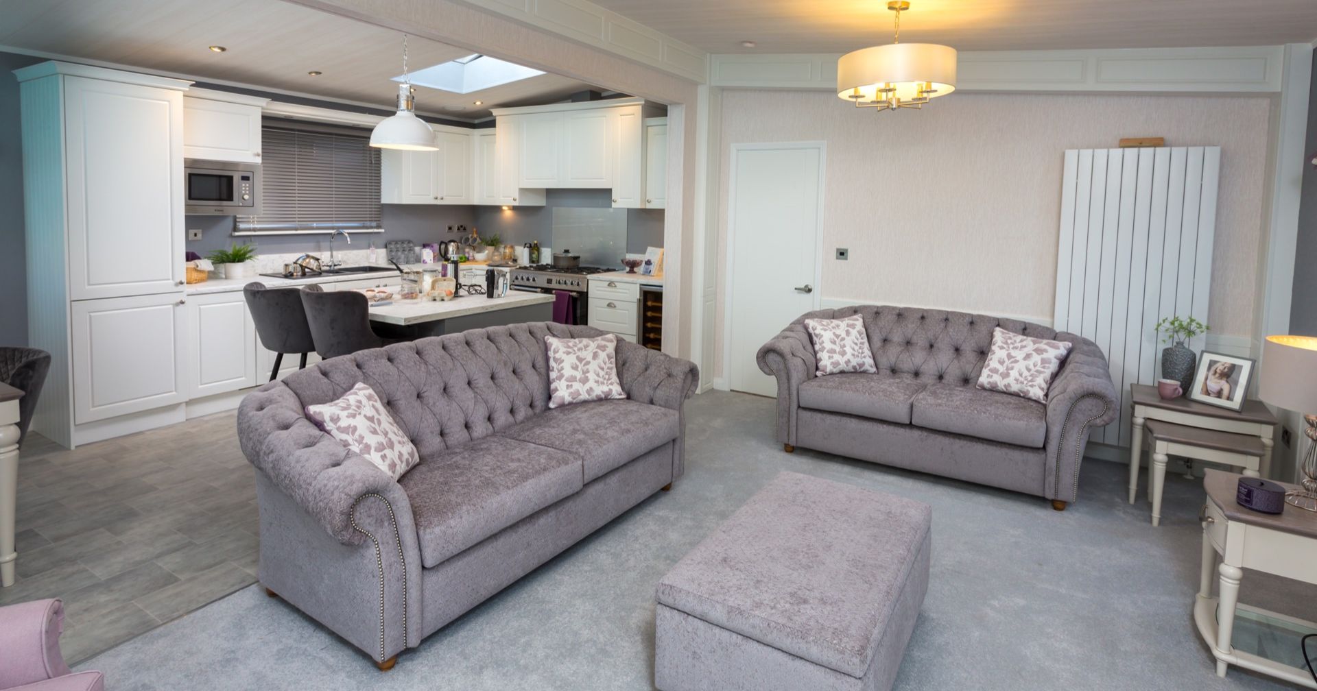 Open-plan living room and kitchen with grey tufted sofas, ottoman, and white cabinetry