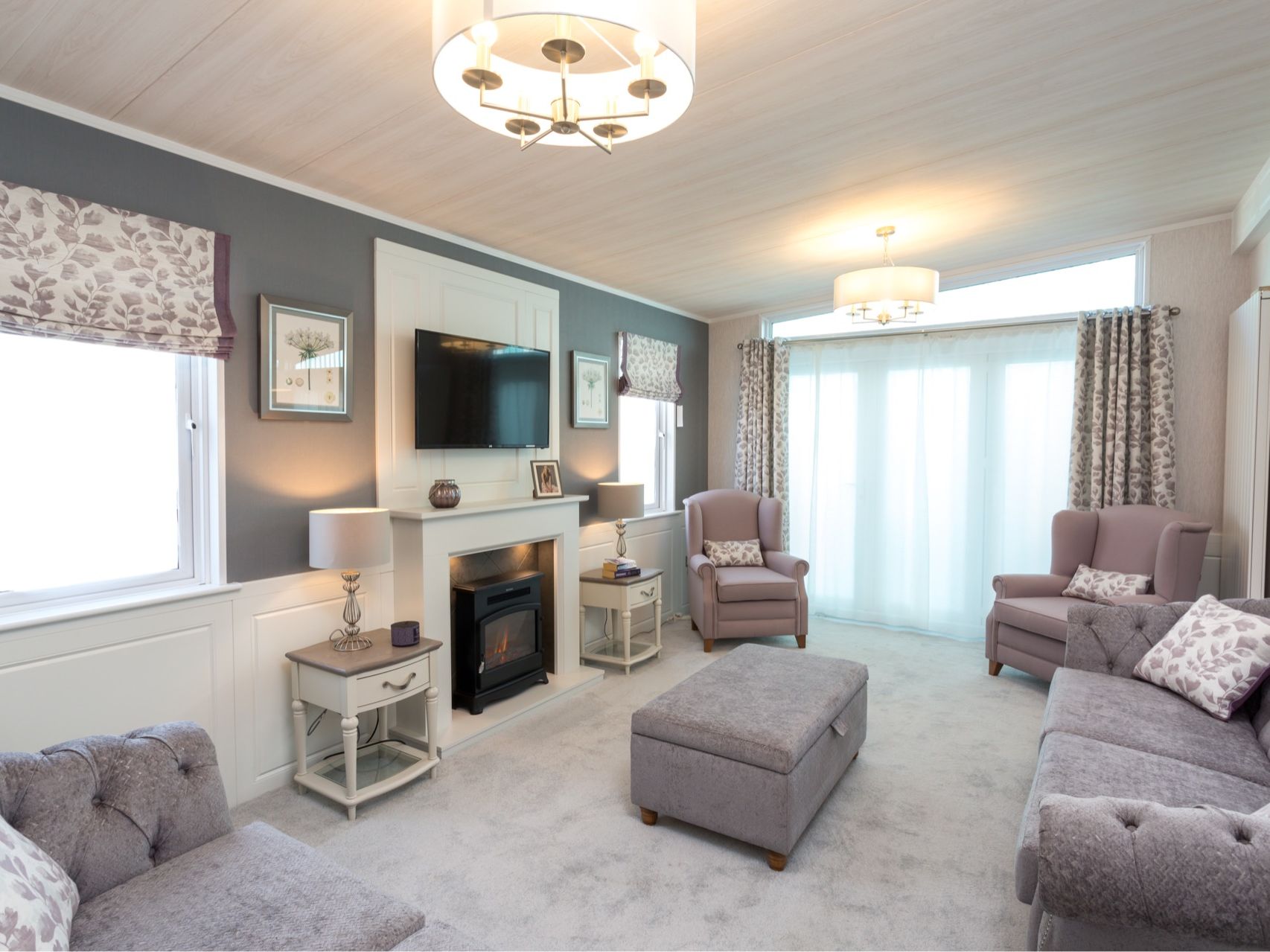 Bright modern living room with grey walls, wall-mounted TV above a fireplace, armchairs, sofas, ottoman and large patio doors with patterned curtains.
