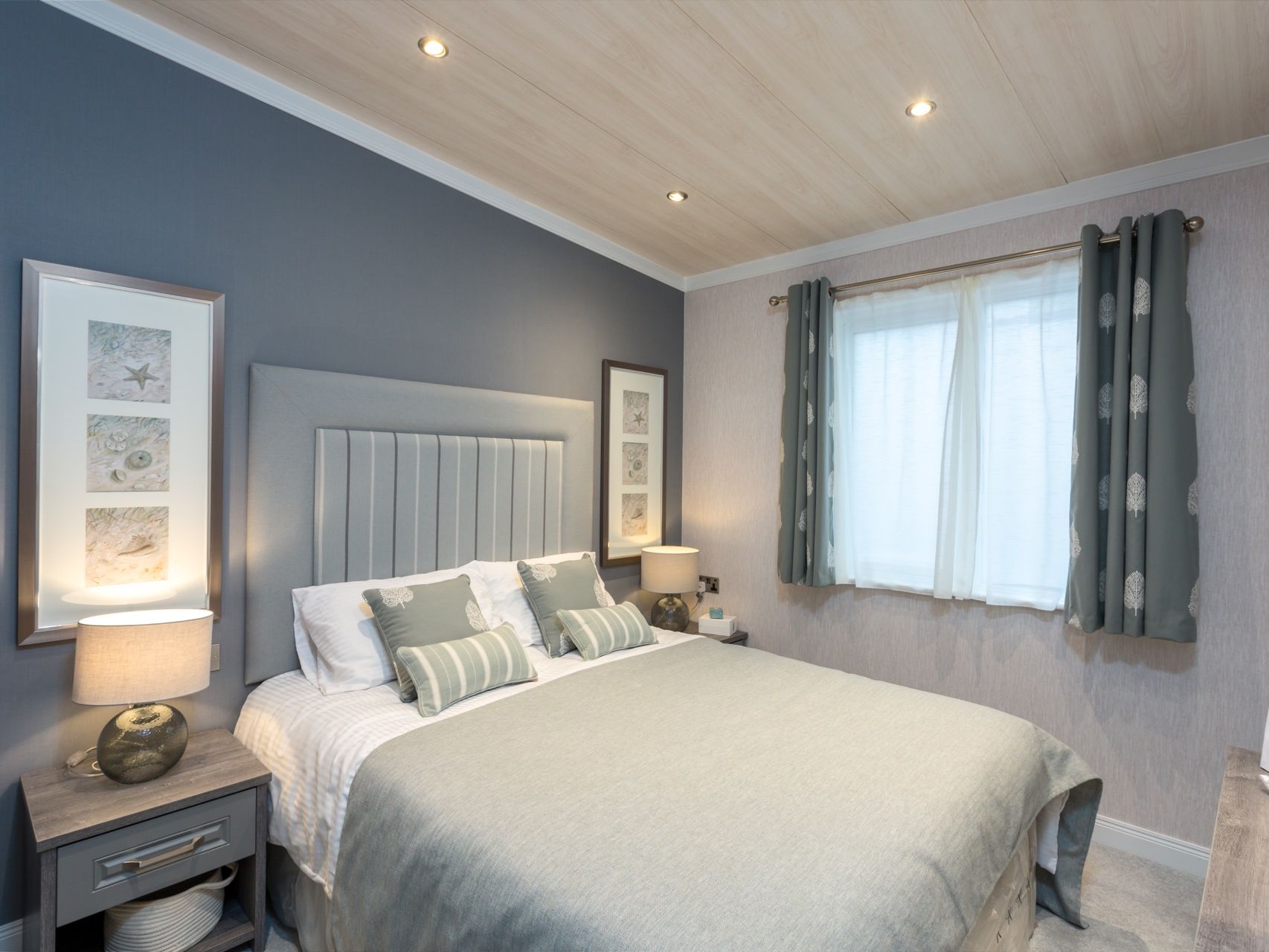 Modern bedroom with grey upholstered headboard, bedside lamps, framed wall art, and a window with patterned curtains