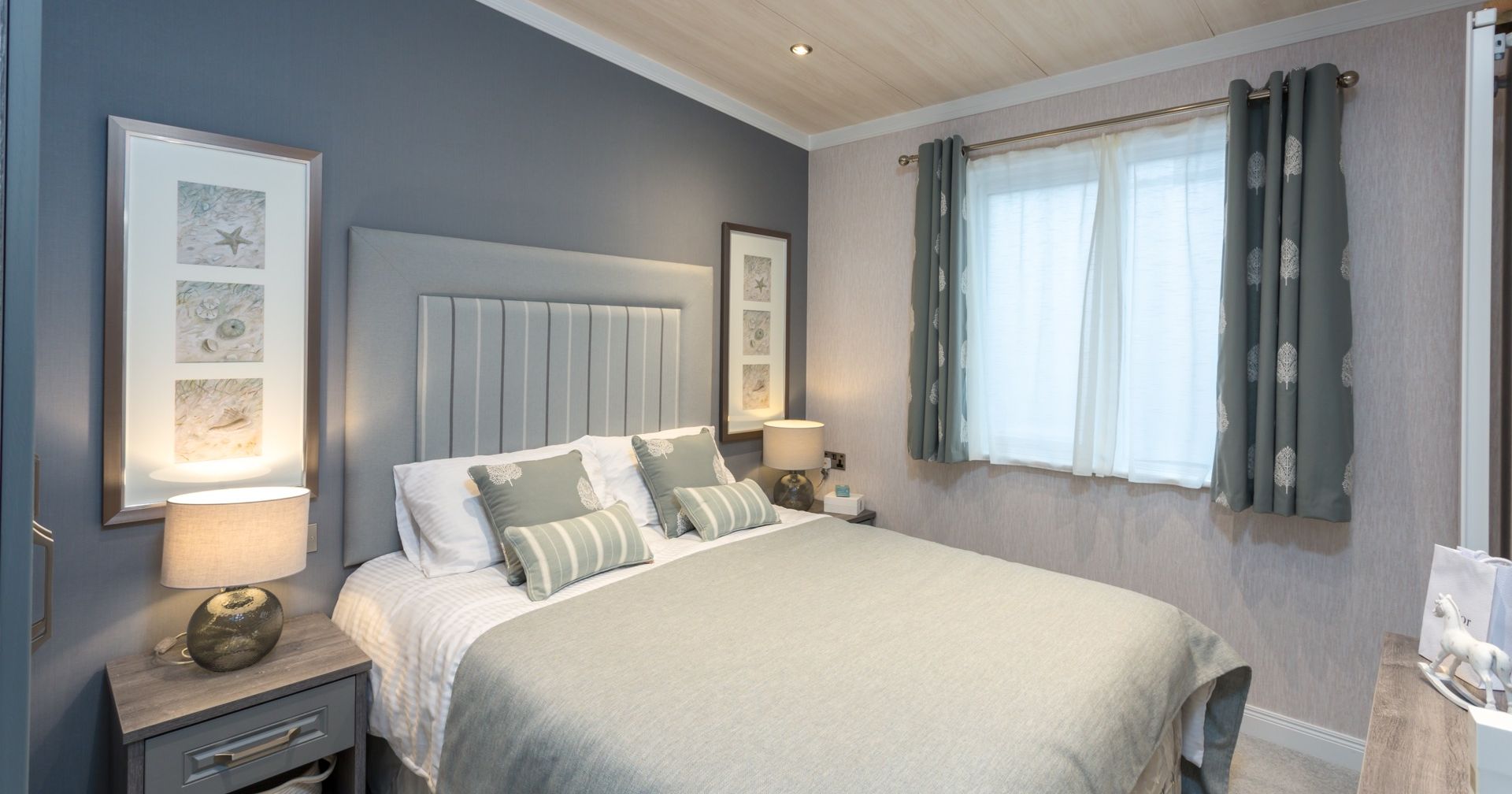 Modern bedroom with grey upholstered headboard, bedside lamps, framed wall art, and a window with patterned curtains