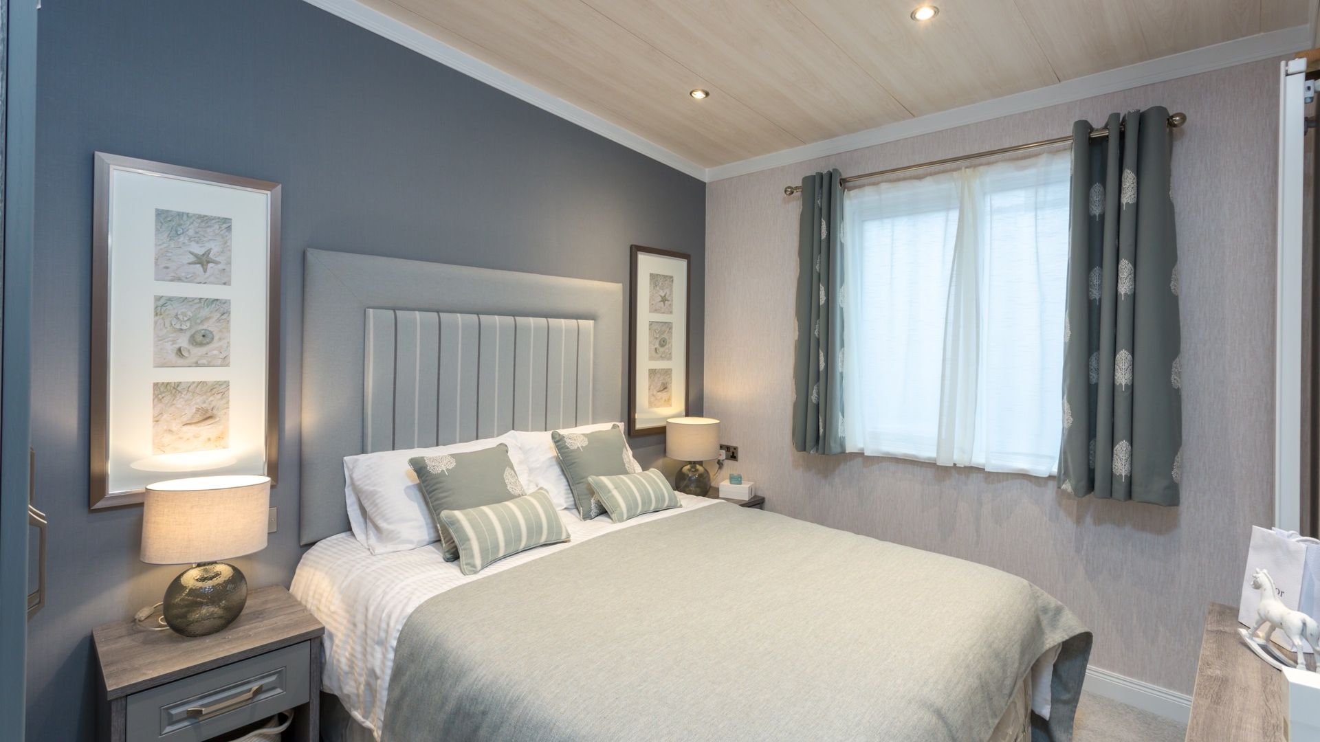Modern bedroom with grey upholstered headboard, bedside lamps, framed wall art, and a window with patterned curtains