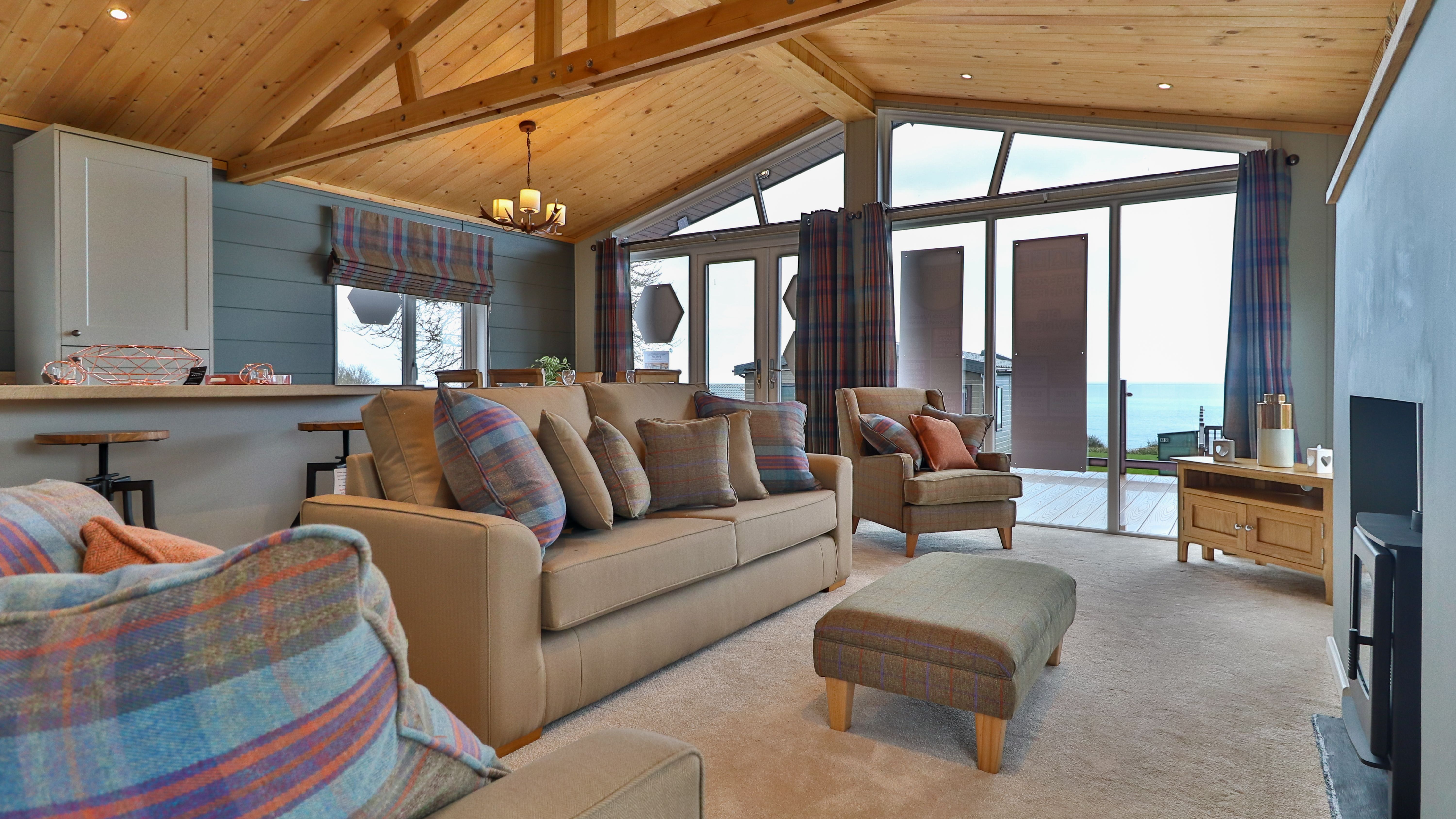 Cosy living room with vaulted wooden ceiling, plaid cushions, and large glass doors opening to a sea view