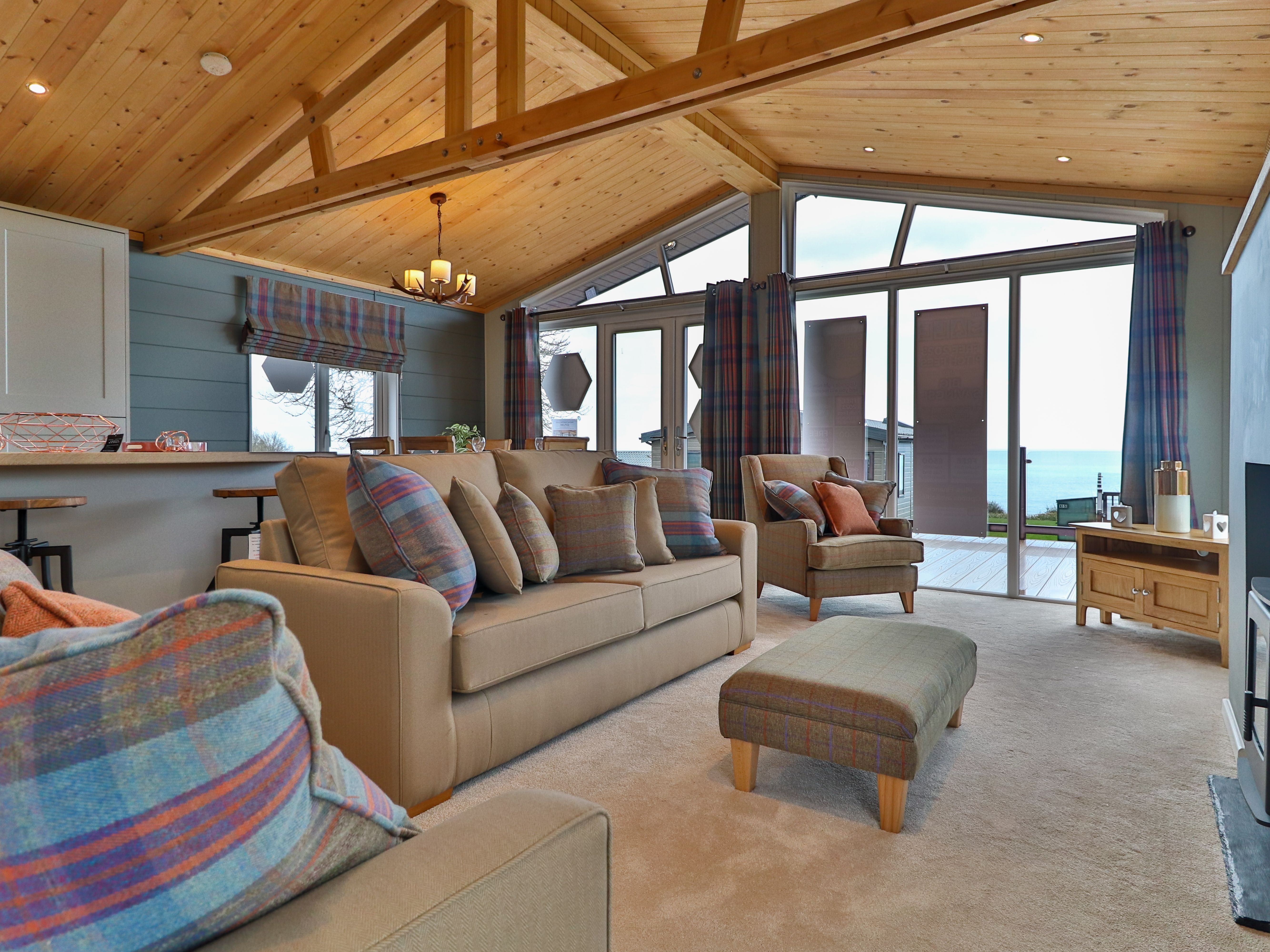 Cosy living room with vaulted wooden ceiling, plaid cushions, and large glass doors opening to a sea view