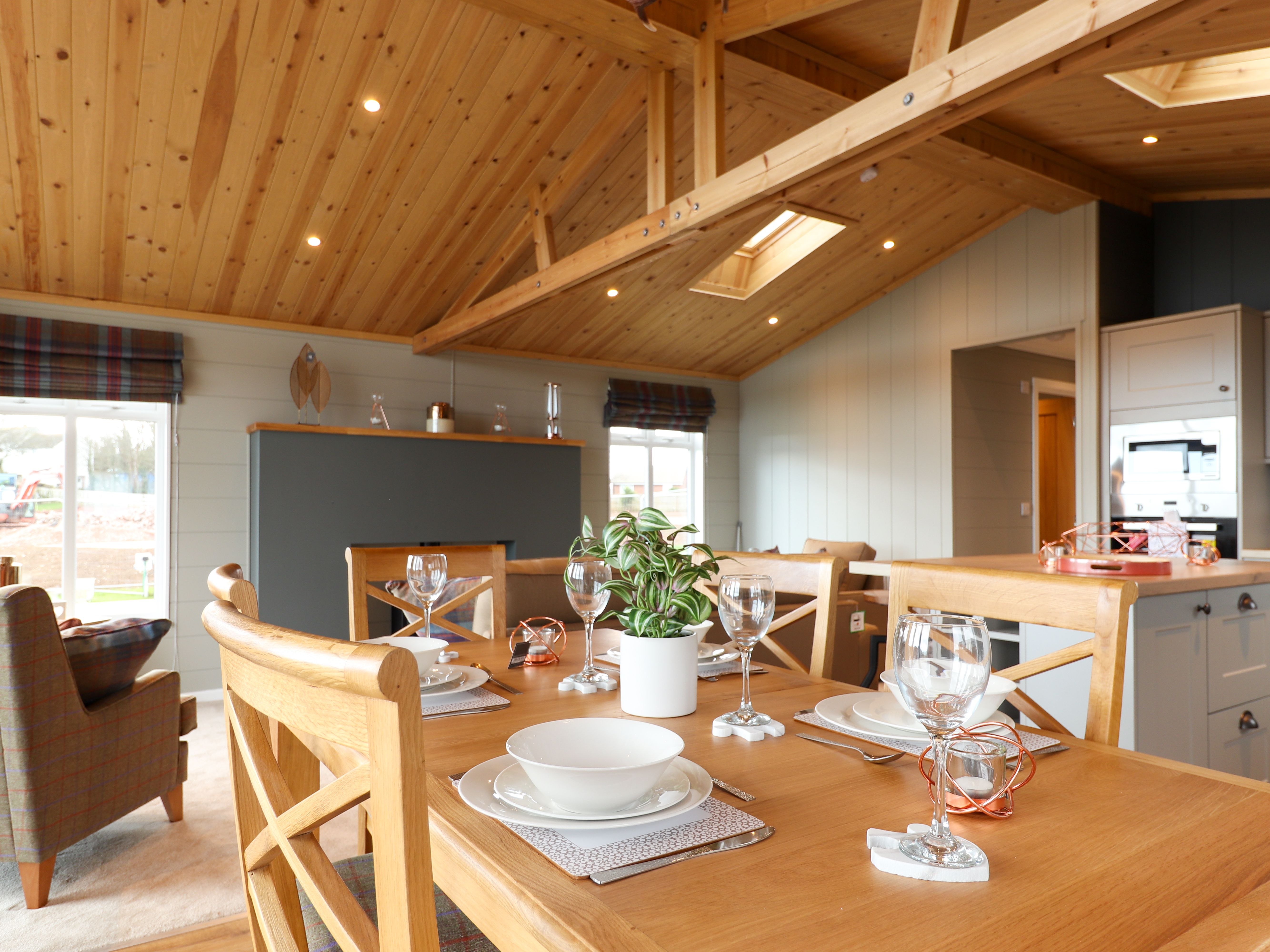 Open-plan dining area with wooden table set for four beneath a vaulted timber ceiling, adjoining a modern kitchen.