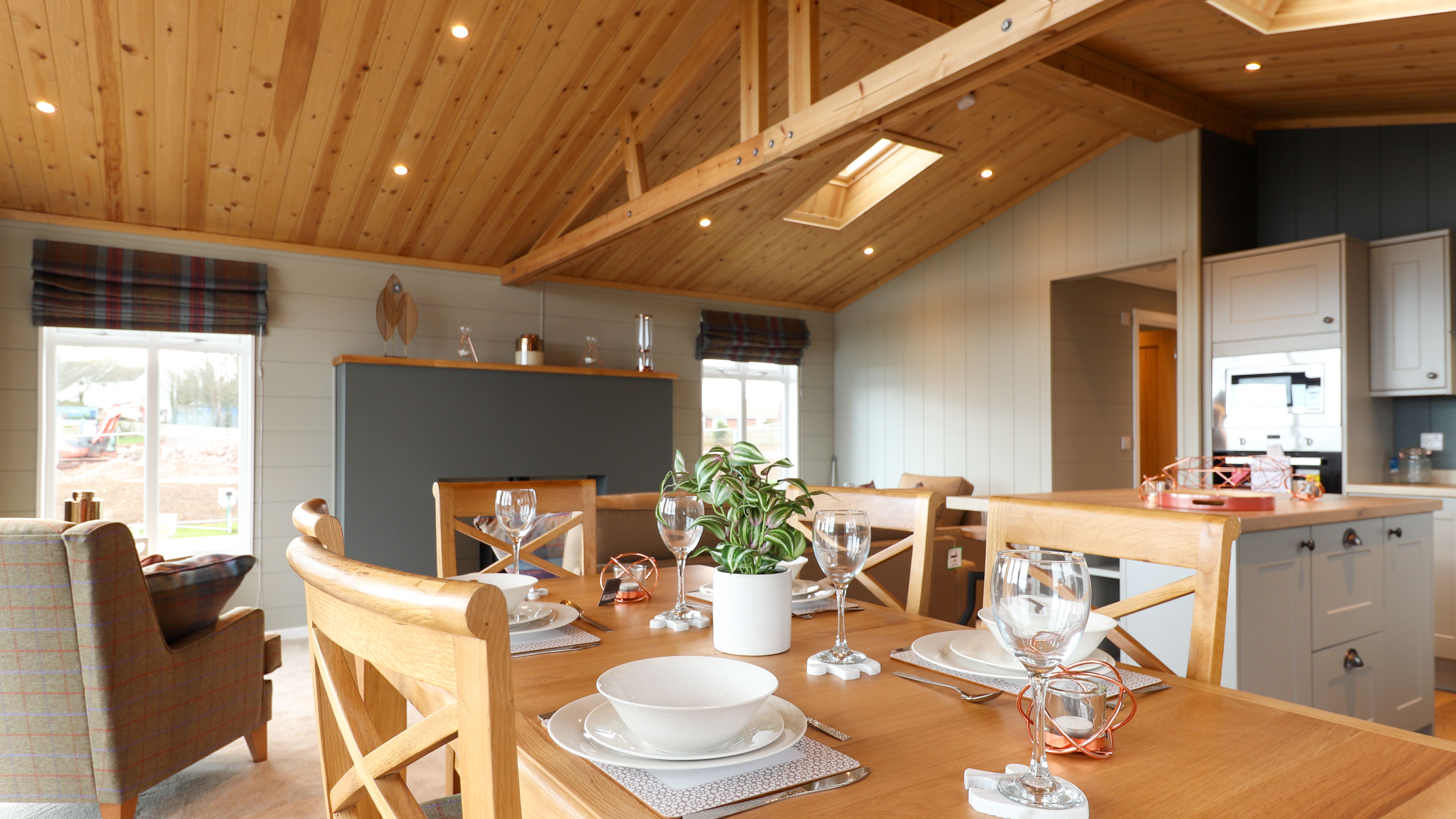 Open-plan dining area with wooden table set for four beneath a vaulted timber ceiling, adjoining a modern kitchen.