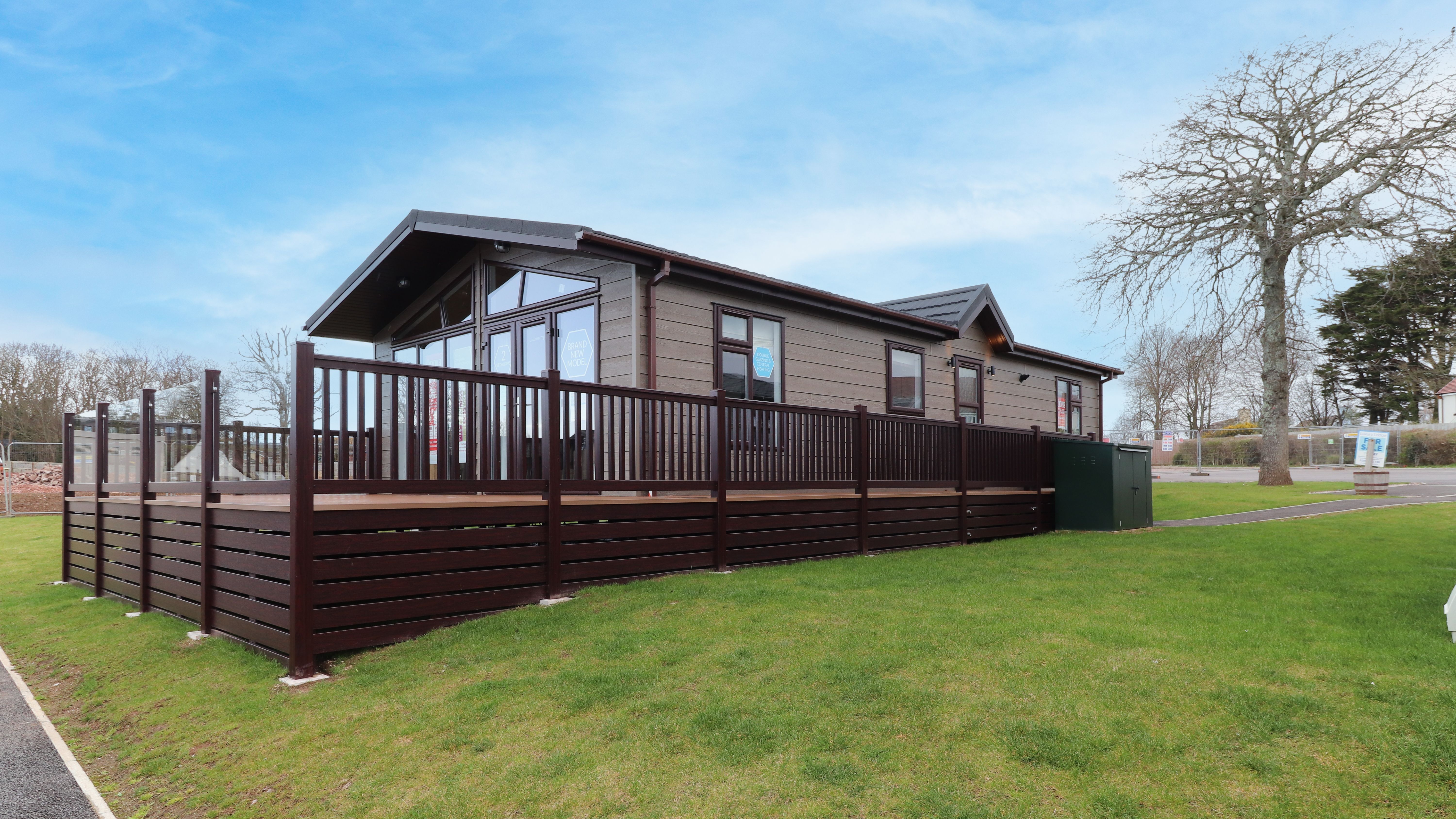 Modern lodge-style caravan with a large wooden deck on a grassy plot under a blue sky