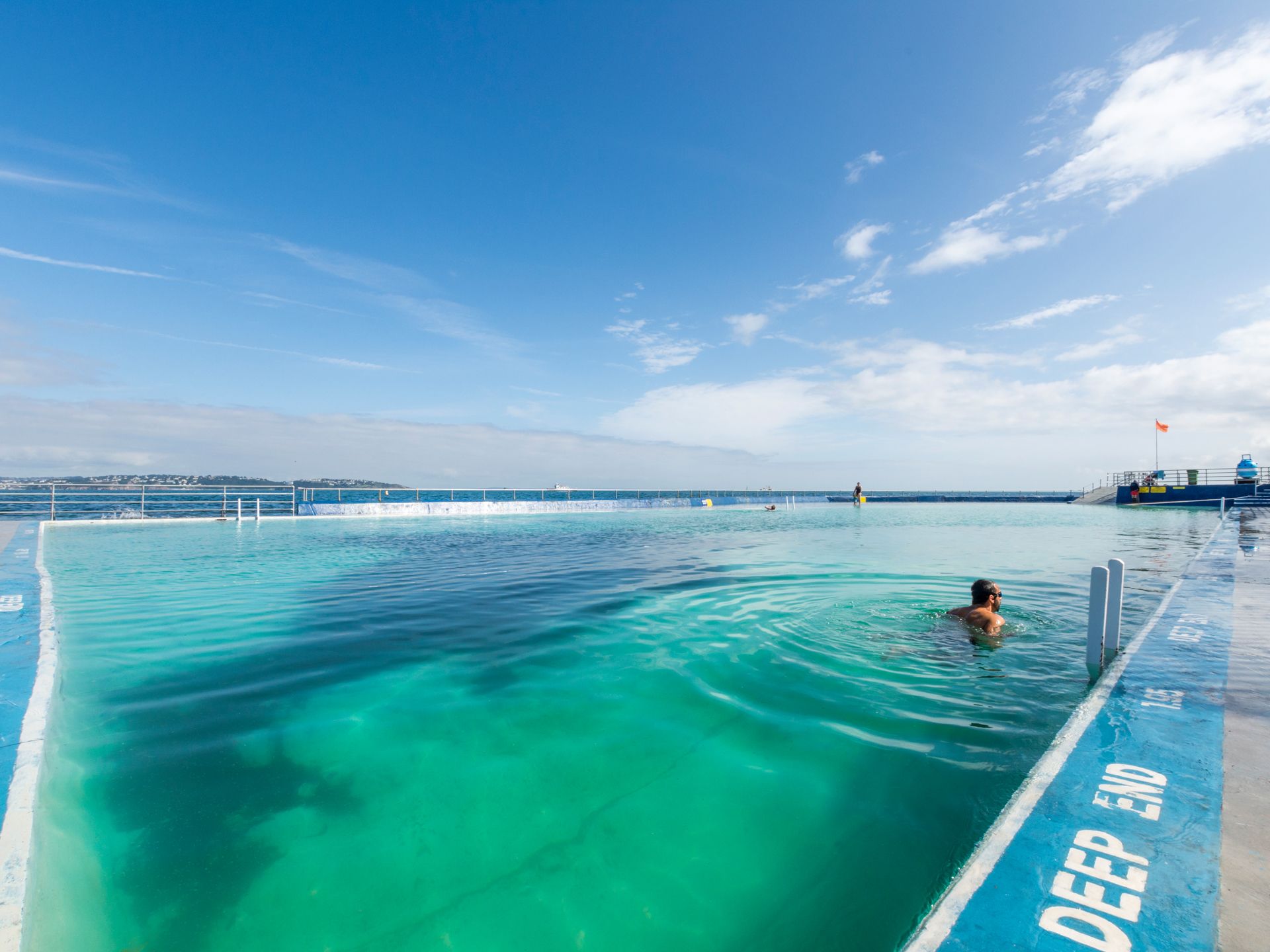 Outdoor ocean pool with turquoise water under a bright blue sky, with a swimmer near the edge