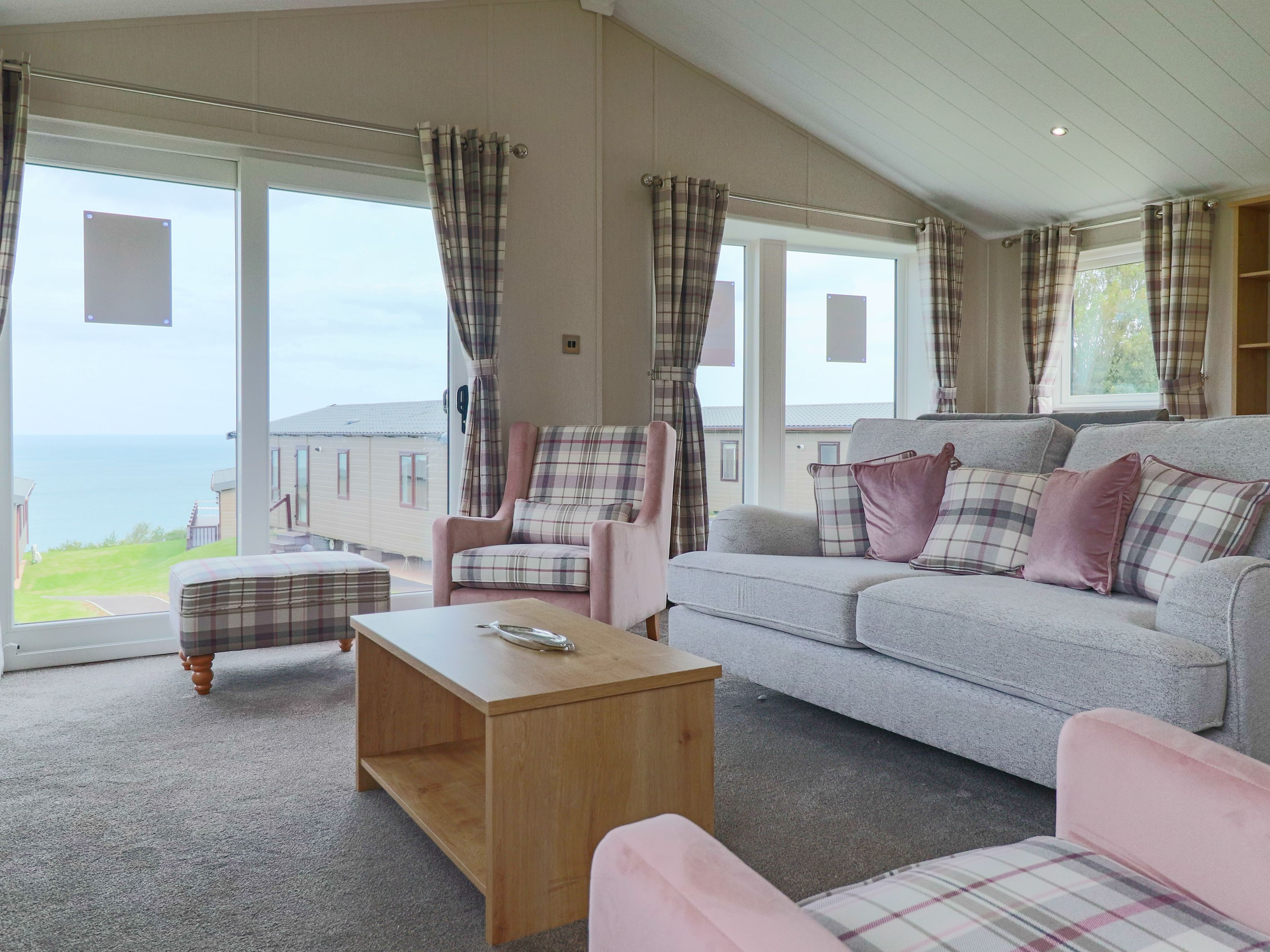 Bright lounge with grey sofa, pink armchairs and plaid curtains beside large windows overlooking the sea