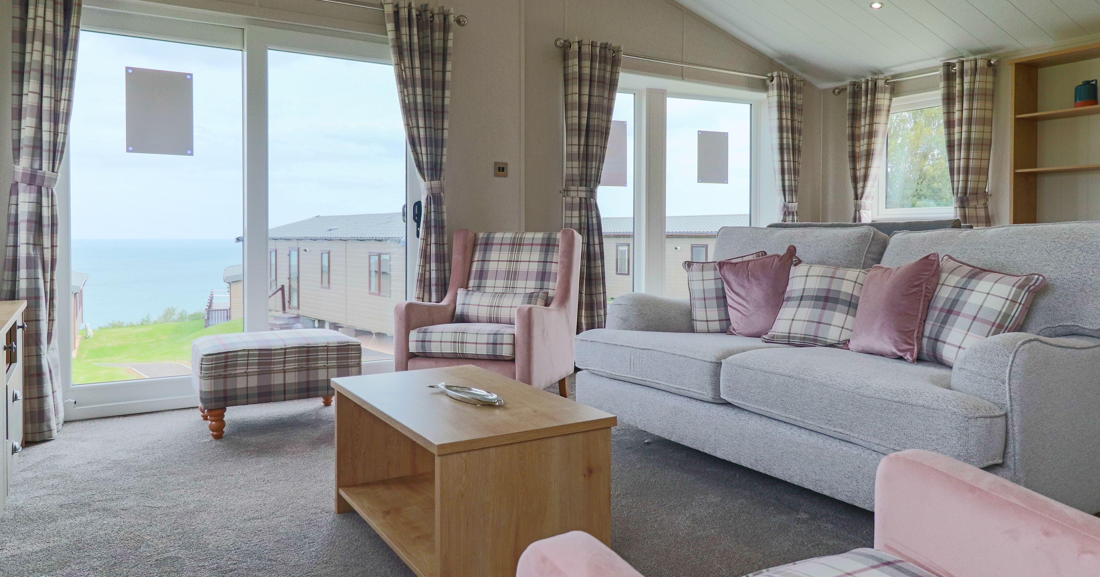 Bright lounge with grey sofa, pink armchairs and plaid curtains beside large windows overlooking the sea