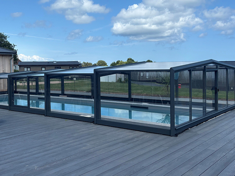 Covered outdoor swimming pool with glass enclosure on a grey deck beside holiday lodges