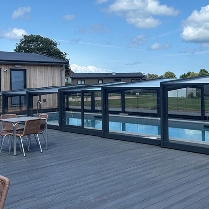 Covered outdoor swimming pool with glass enclosure on a grey deck beside holiday lodges