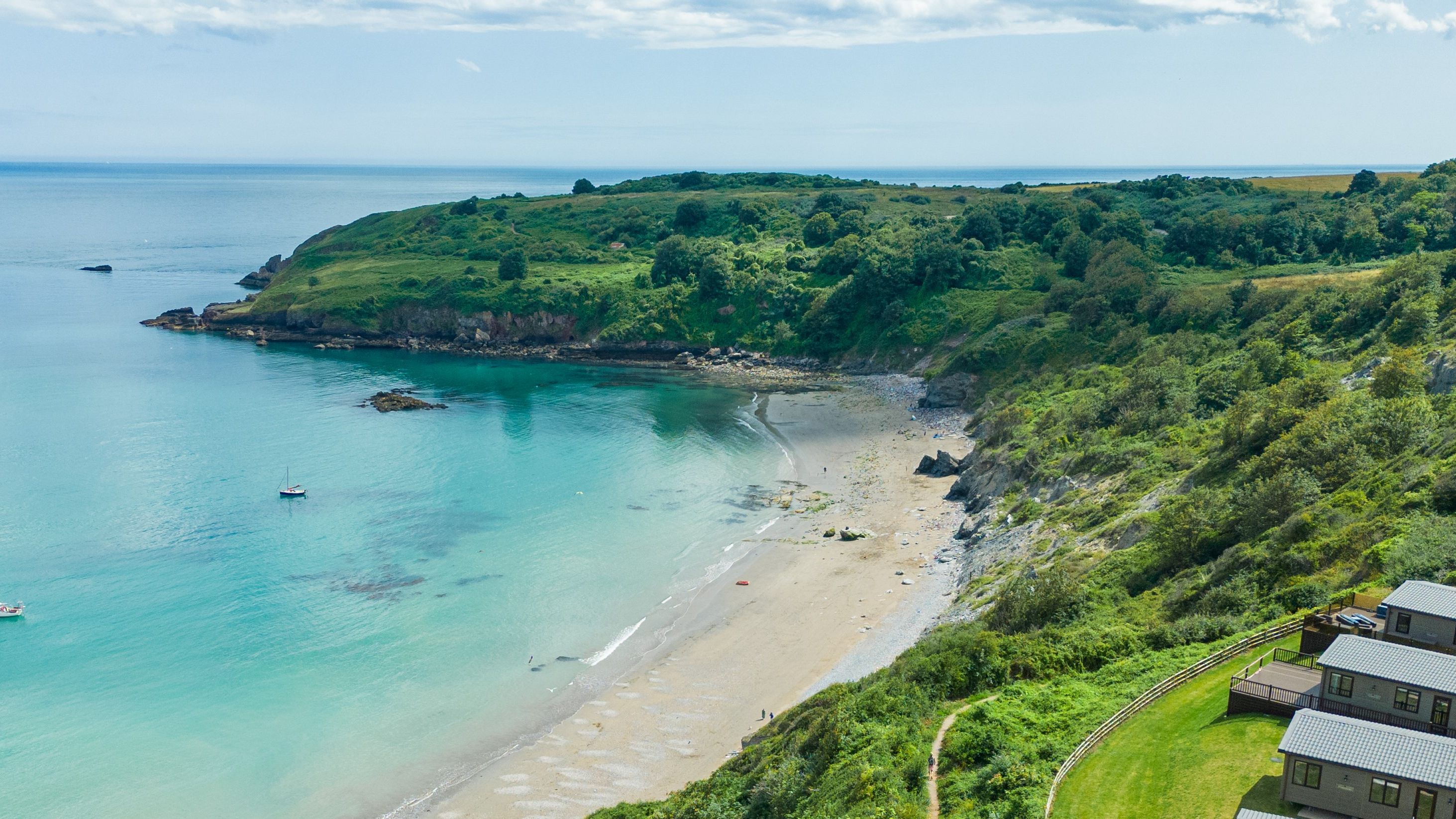 Aerial view of a sandy bay with turquoise sea, green cliffs and a few small boats, with holiday lodges on the hillside