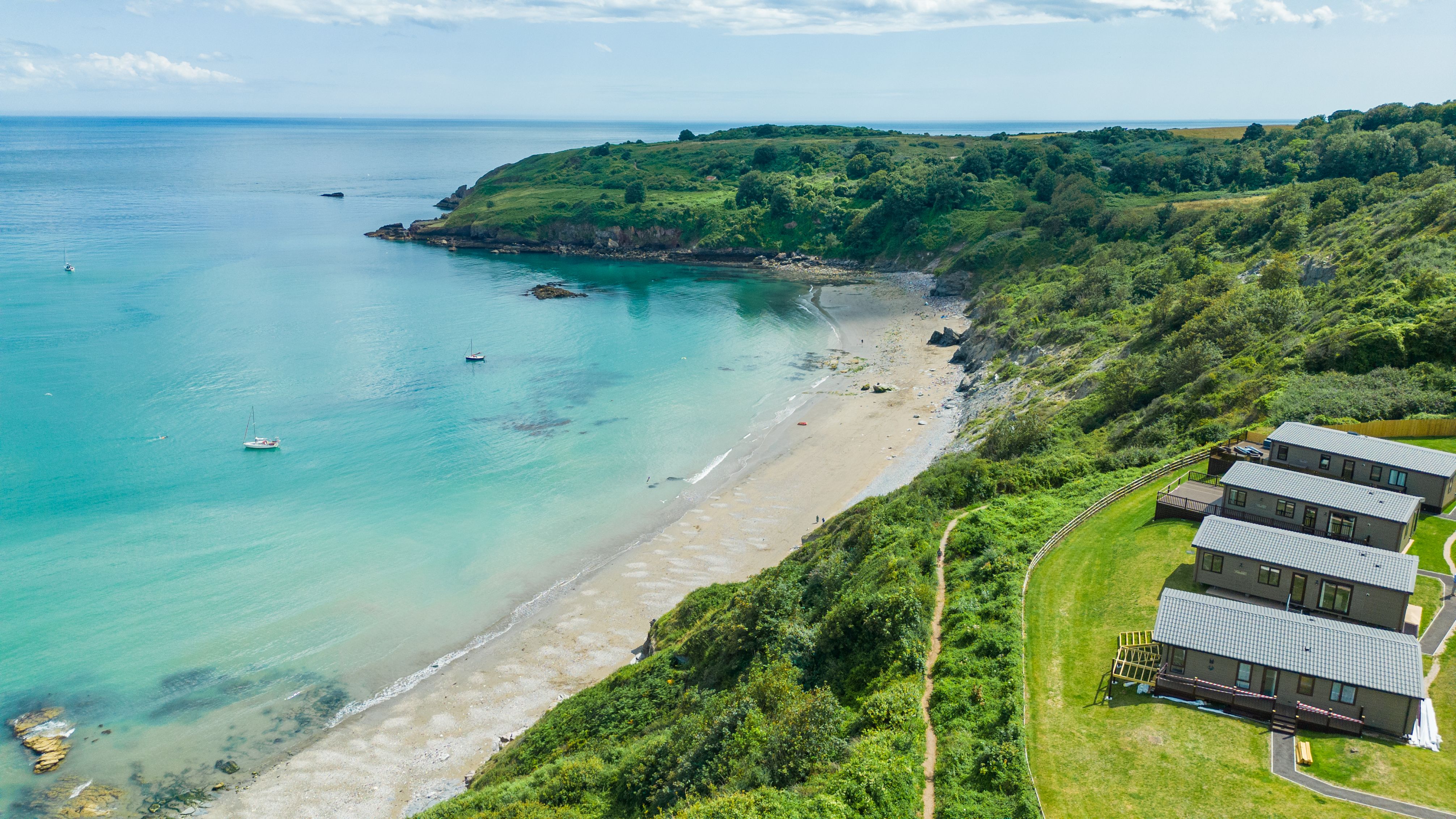 Aerial view of a sheltered sandy bay with turquoise sea, green cliffs and holiday lodges on the hillside