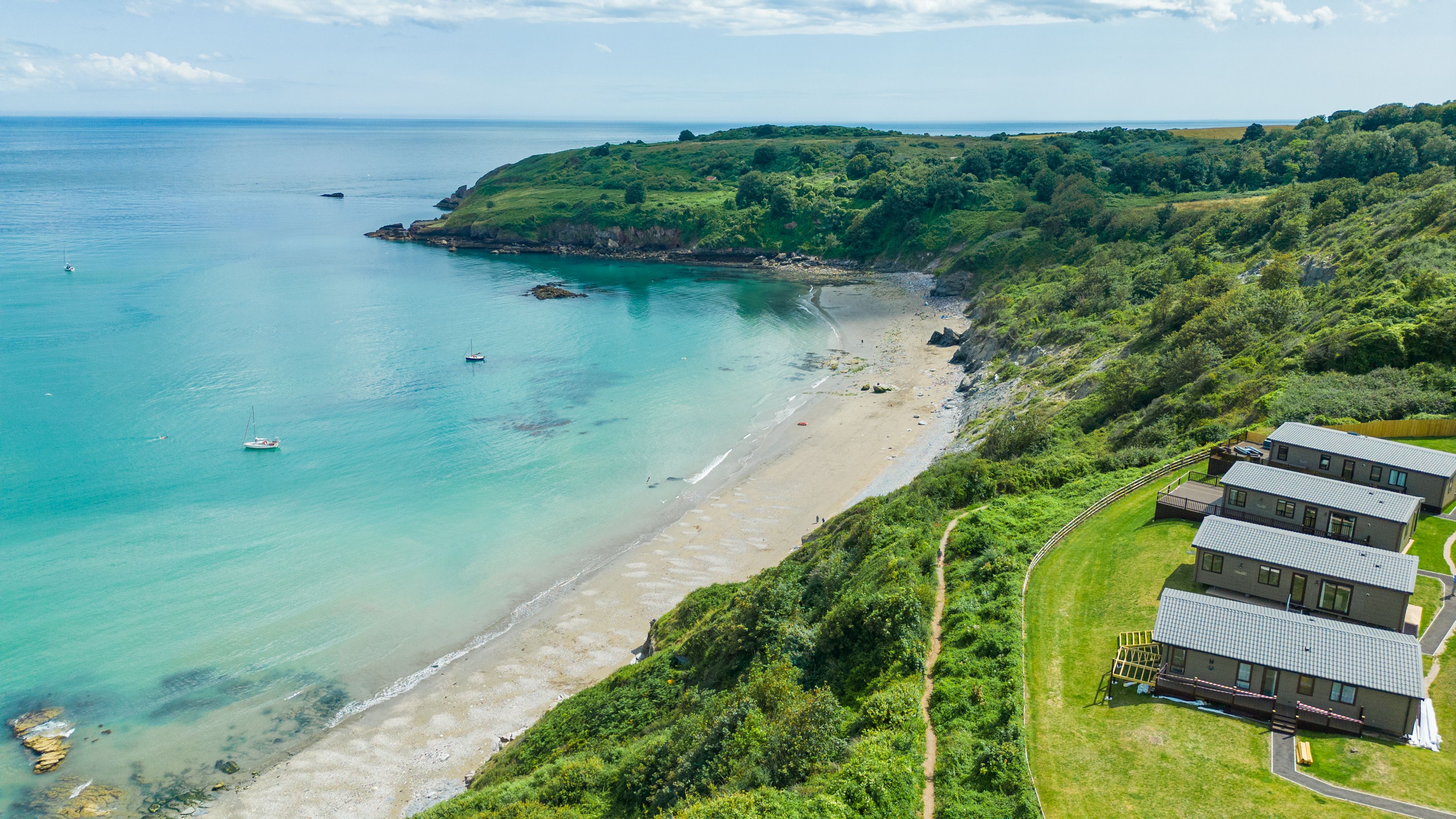 Aerial view of a sheltered sandy bay with turquoise sea, green cliffs and holiday lodges on the hillside